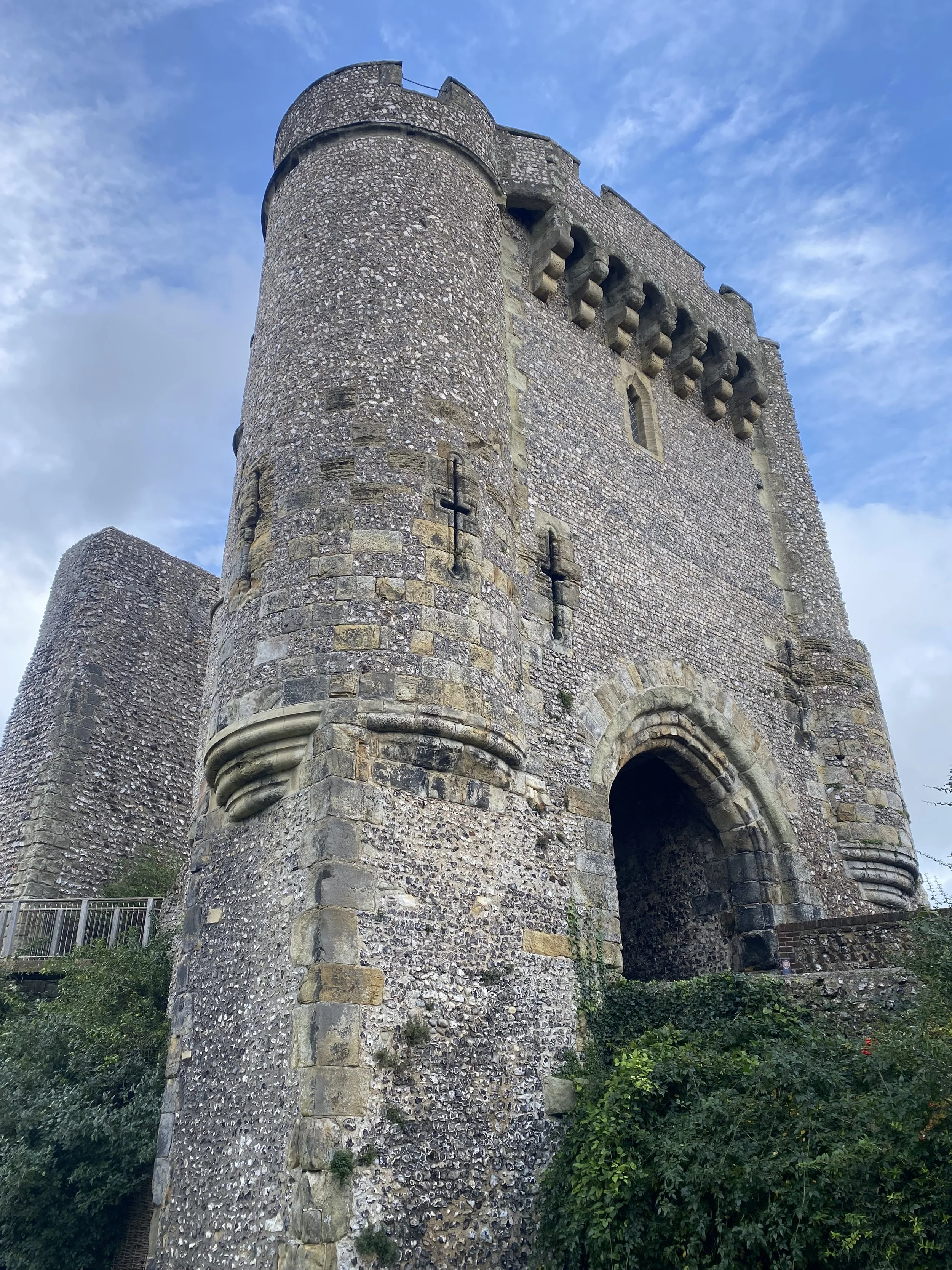 A tall stone medieval castle tower with a large archway at the bottom and small narrow windows near the top, with a backdrop of blue sky with clouds and green foliage at the base.