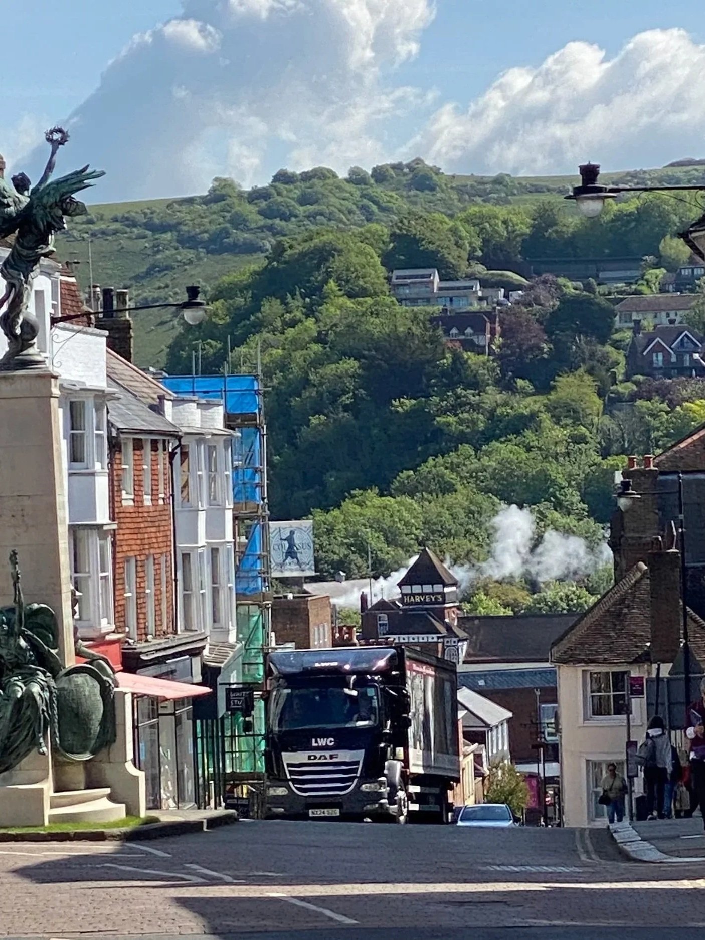 Street view with buildings, a black truck labeled DAF and LWC, hanging street lamps, a sign for Harveys, and hills with lush green trees and houses in the background.