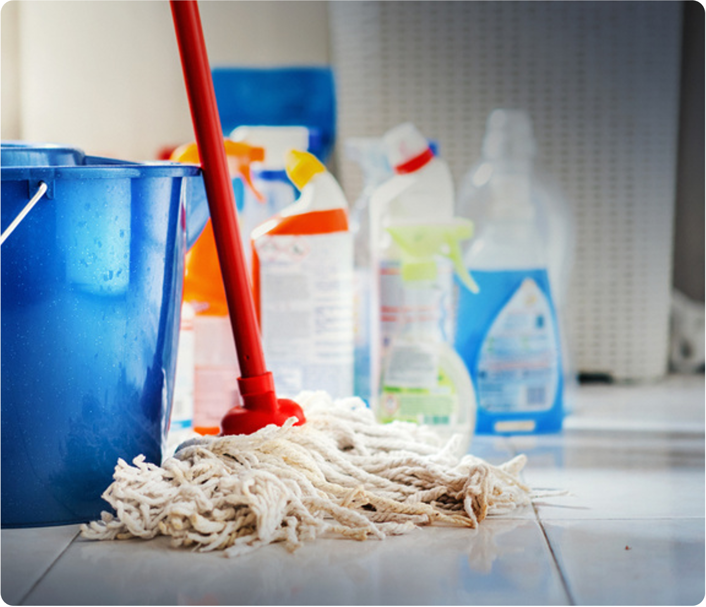 Cleaning mop with red handle and tangles of white cleaning cloth on a tiled floor, with cleaning products in bottles on a shelf in the background.