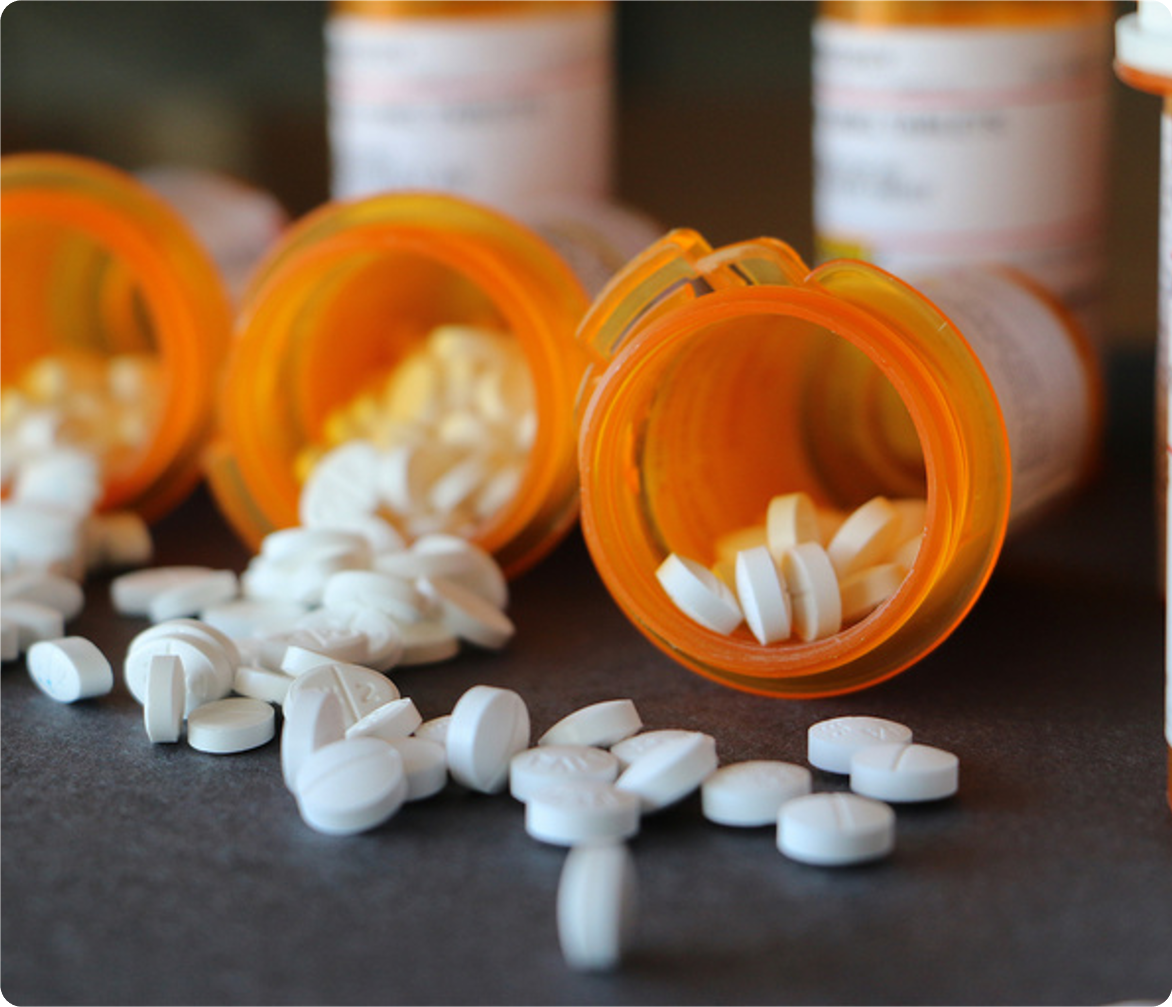 Close-up of scattered white round pills and an orange prescription pill bottle on a dark surface, with several more bottles blurred in the background.