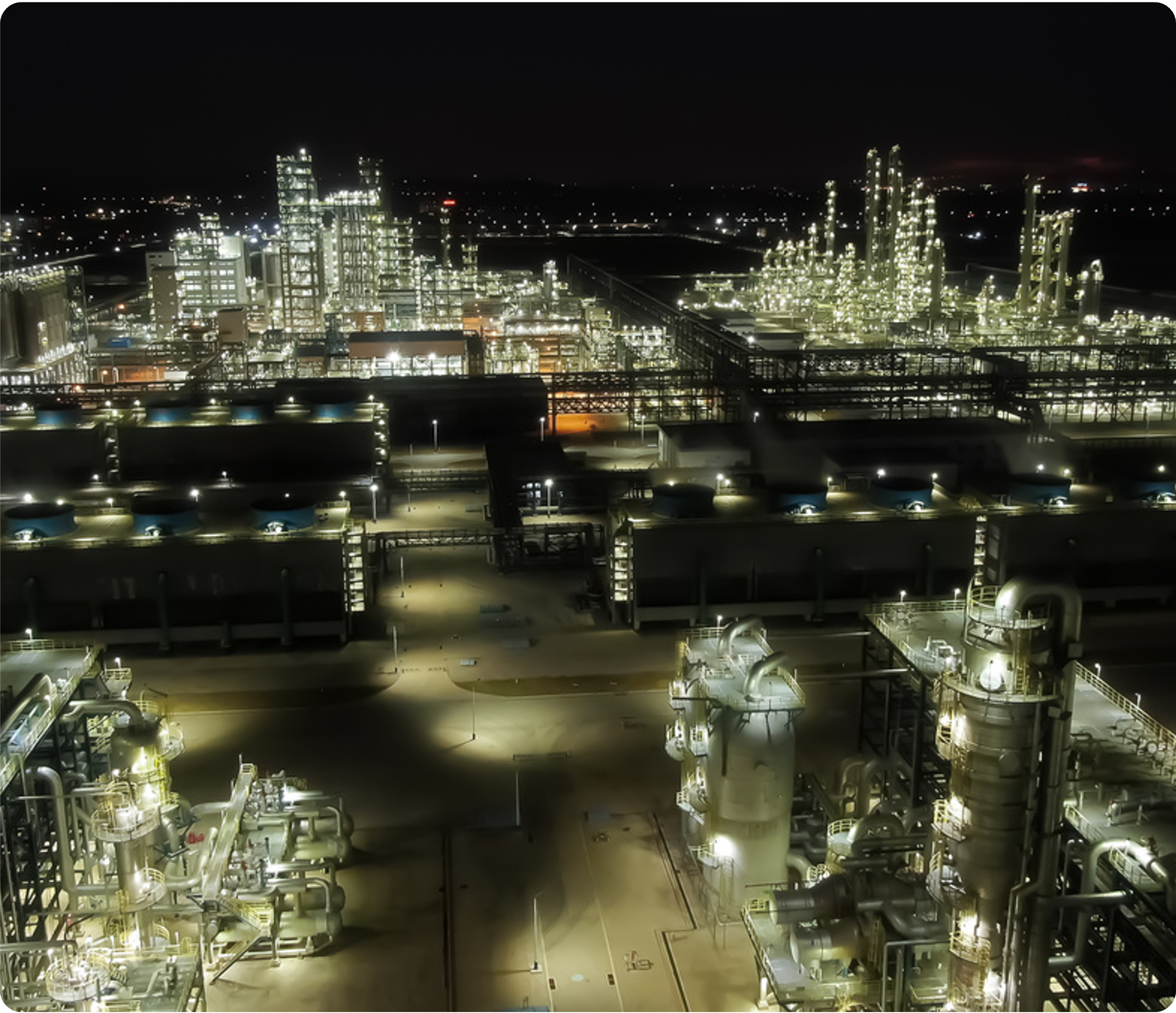 Night view of an industrial plant illuminated with numerous bright lights, featuring pipelines, towers, and structures, with a dark sky in the background.