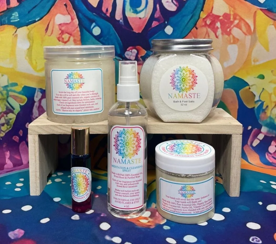 Collection of Namaste bath and body products including bath salts, a spray, a massage or cleansing oil, and body butter on a wooden platform with colorful abstract art in the background.