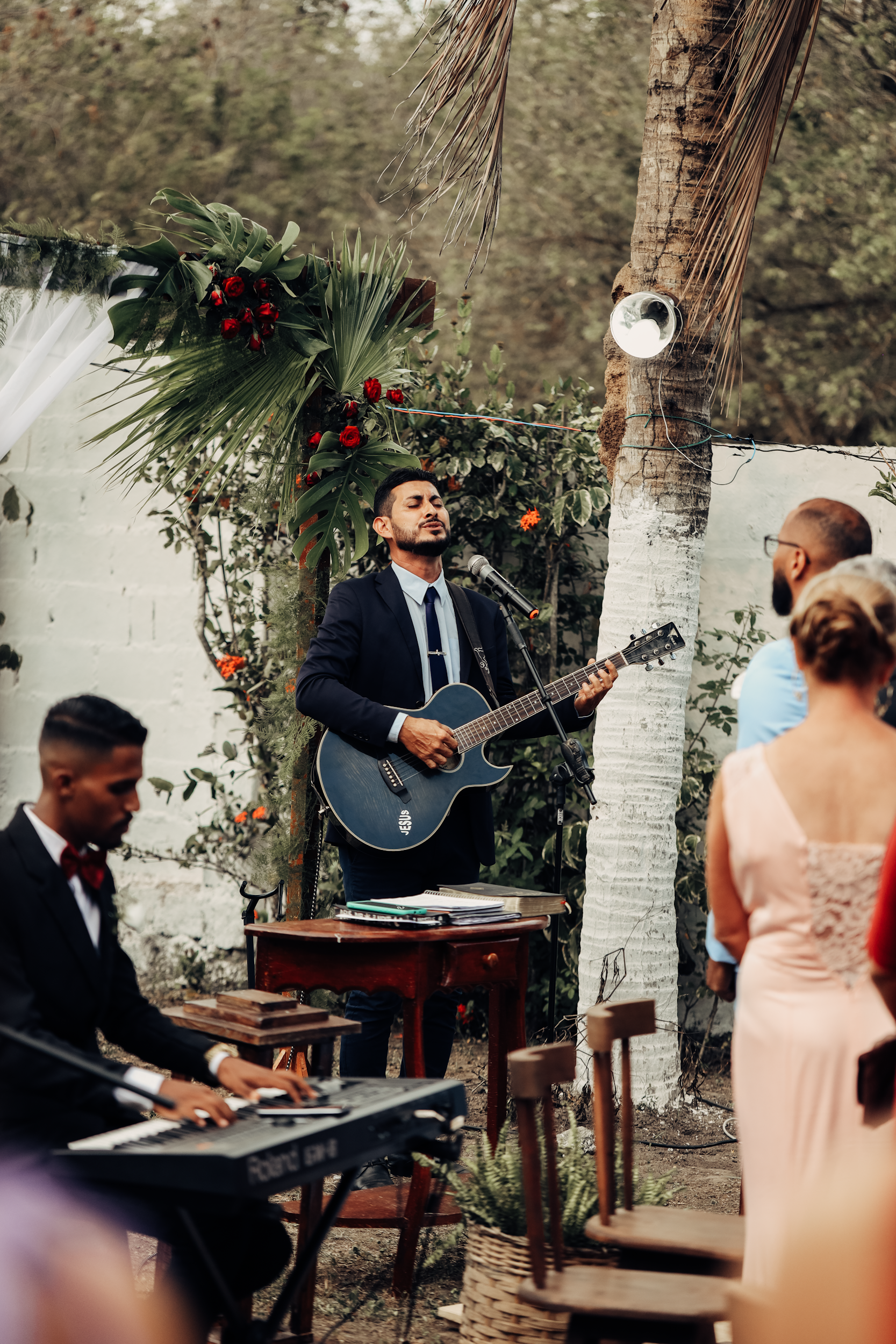 A man in a dark suit with a tie and beard is singing and playing guitar during an outdoor event. Another man in a dark suit is playing keyboard, while a woman in a pink dress is standing, facing the performers. The scene is decorated with green plants, palm leaves, and red flowers, with a white wall and palm tree in the background.
