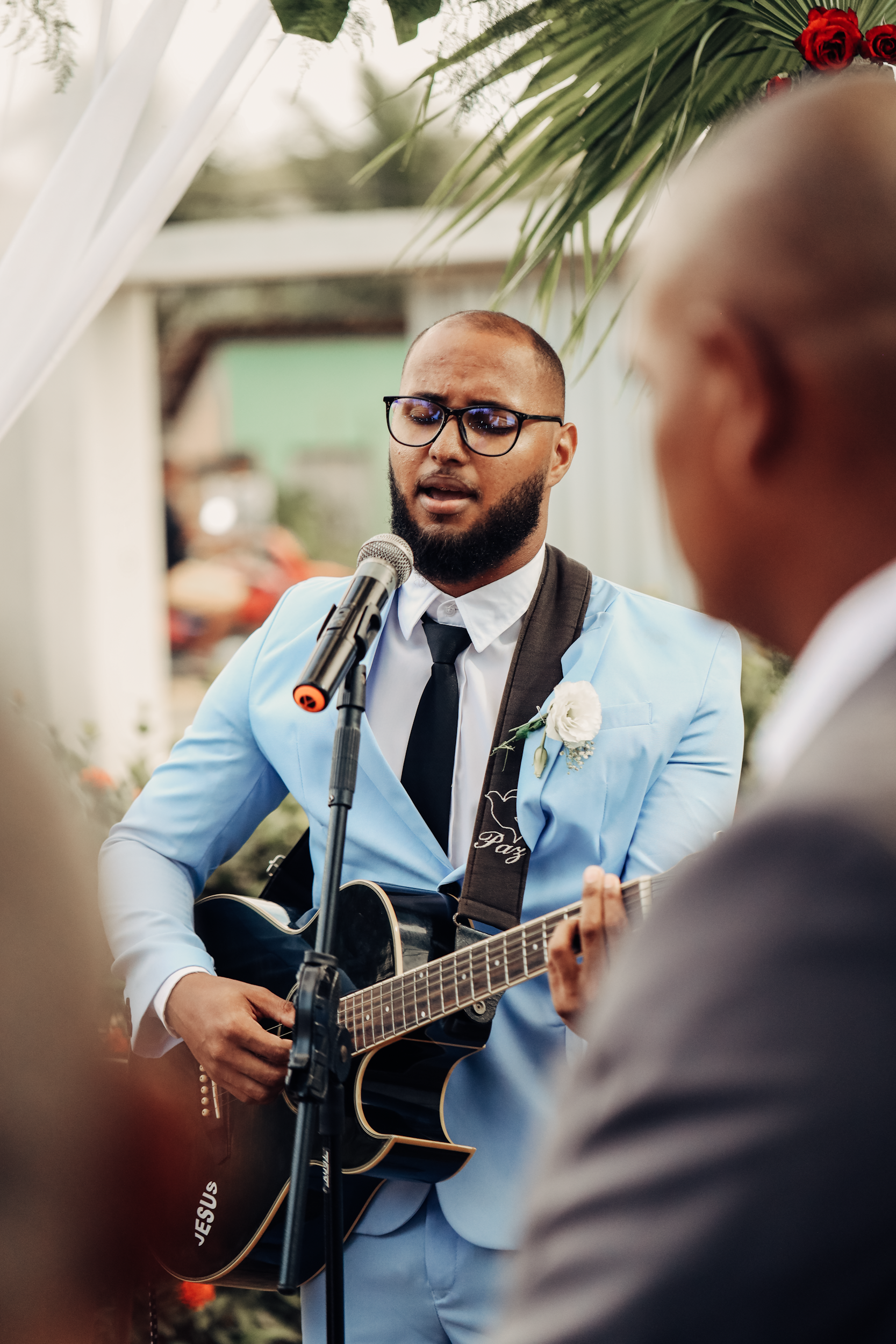 A man with glasses, a beard, wearing a light blue suit and white shirt, is singing into a microphone while playing an acoustic guitar. He has a white boutonniere on his lapel. There is a person in the foreground with a bald head, and some decorative greenery and red flowers are overhead.