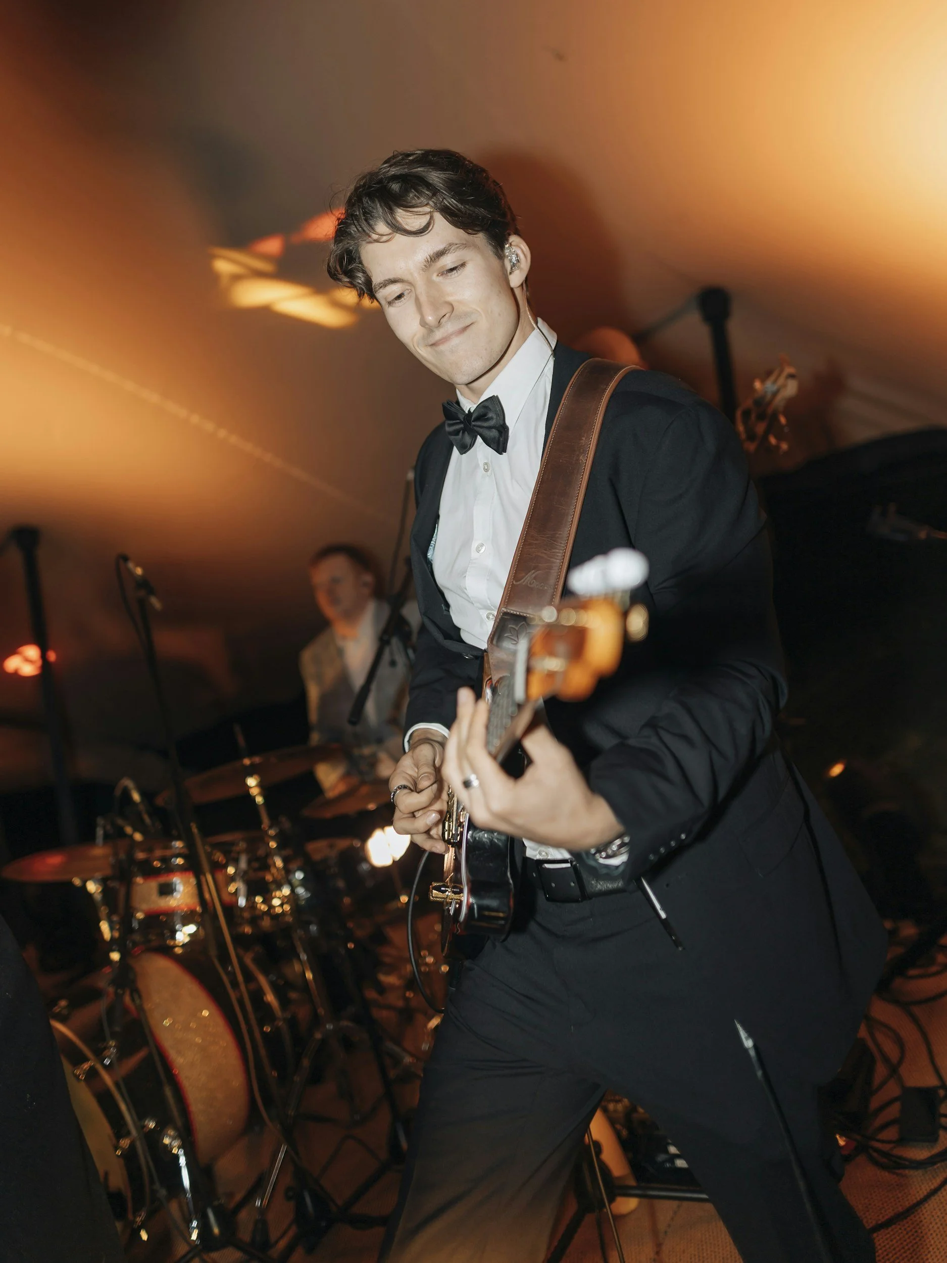 Young man in formal attire playing an electric guitar at a live music event with drums and other musicians in the background, under warm stage lighting.
