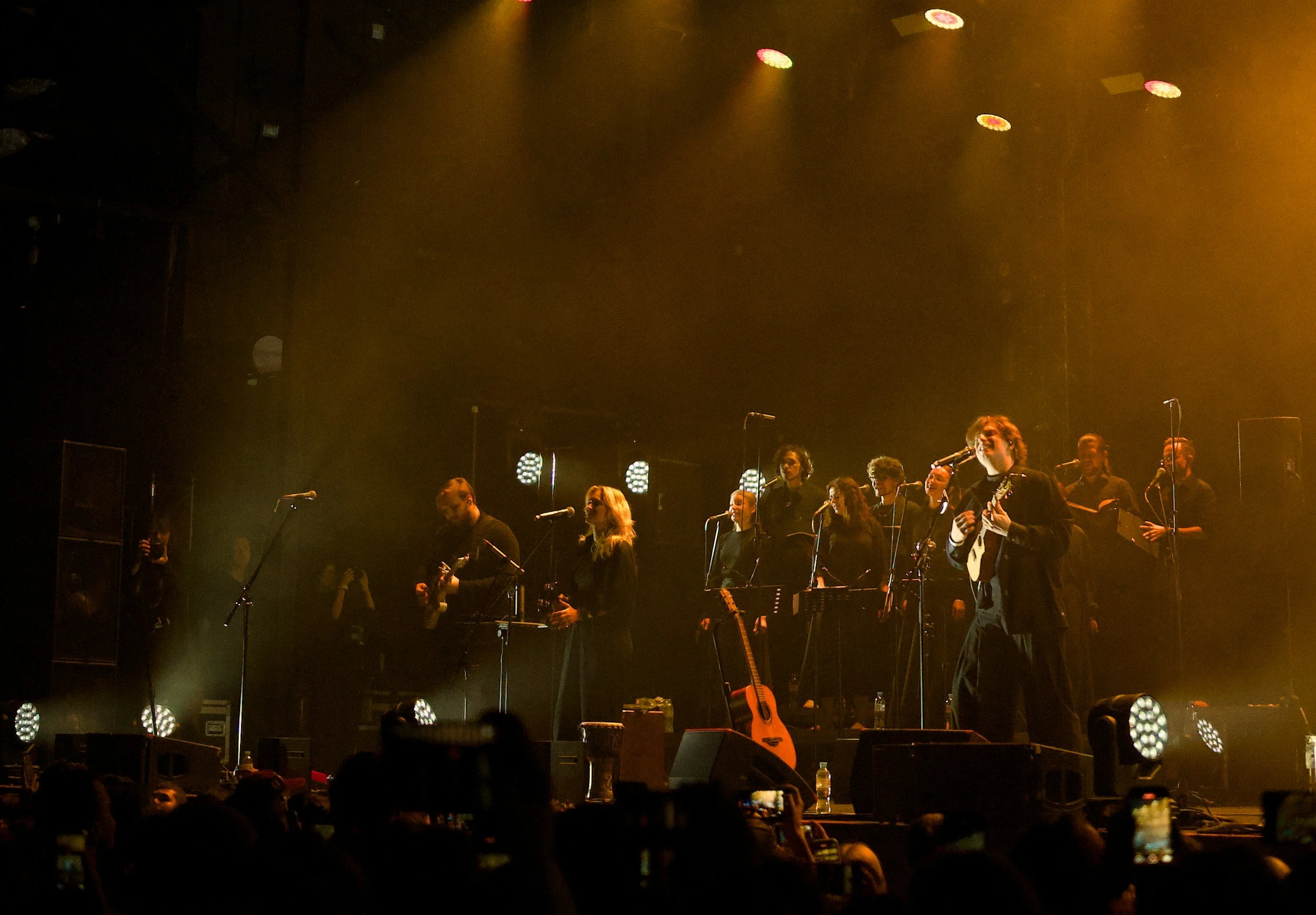 A group of musicians on stage performing live, one woman playing guitar are illuminated by stage lights in a dark concert venue.