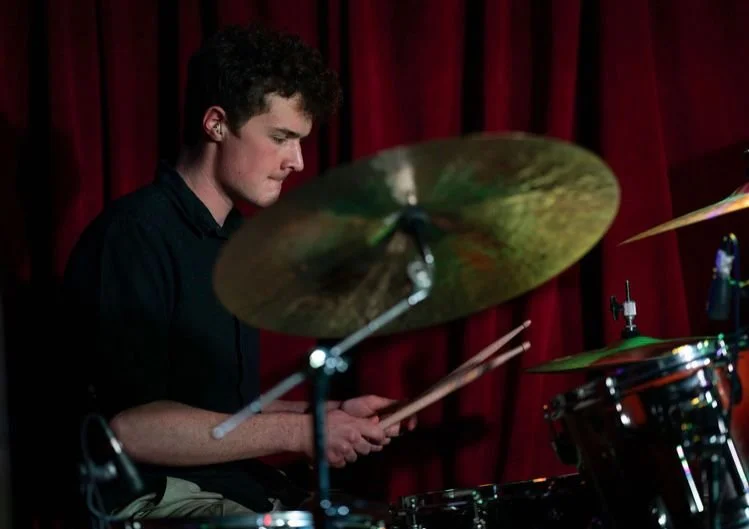 A young man playing drums on stage with red curtains in the background.