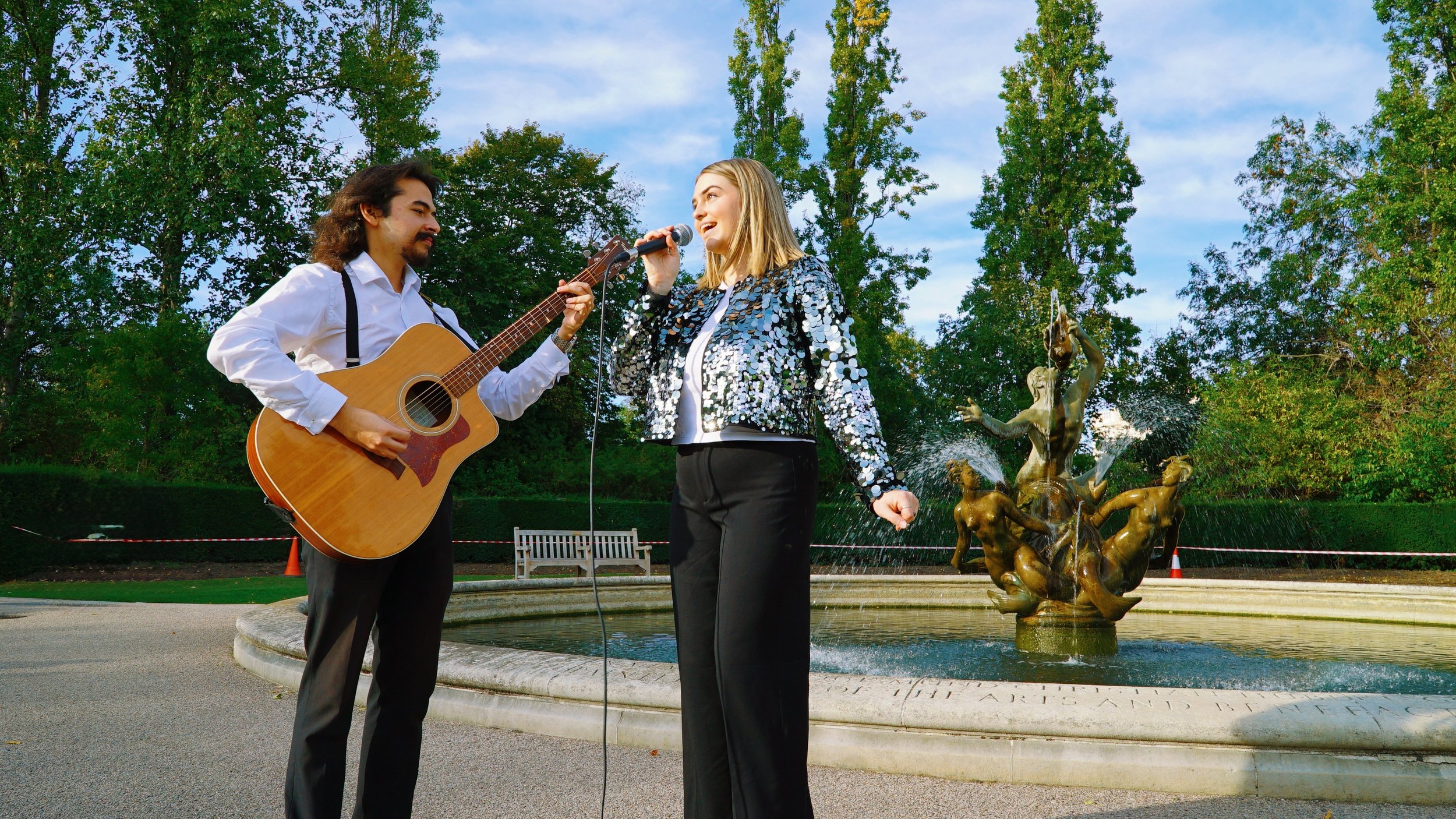 A man and woman performing music outdoors near a fountain in a park, with trees and sky in the background.