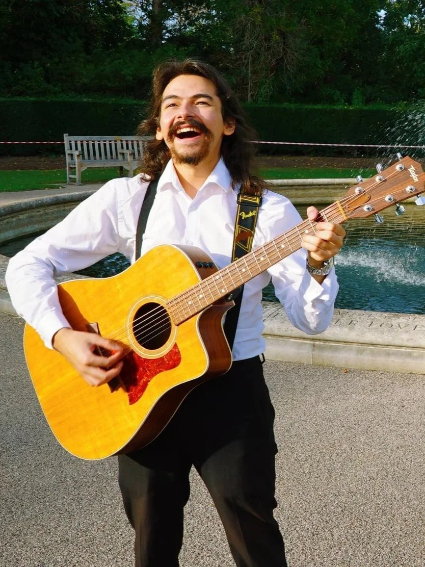 A man with long dark hair and a beard, wearing a white shirt and black pants, is playing an acoustic guitar and smiling, standing outdoors near a fountain with trees in the background.