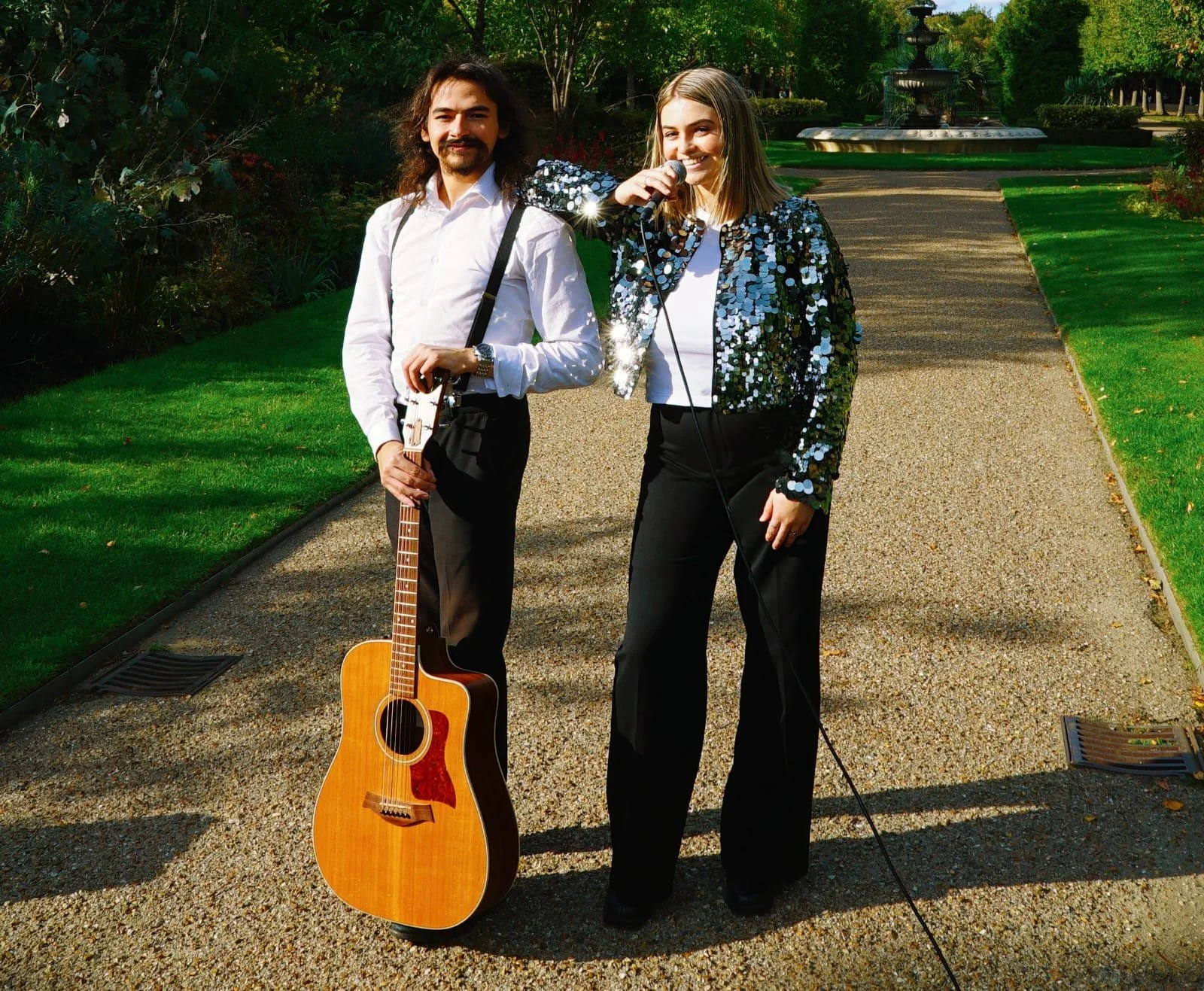 A man with long hair and a beard holding a guitar, standing next to a smiling woman singing into a microphone, both dressed in shiny, sequined jackets, in a park with green grass and trees.