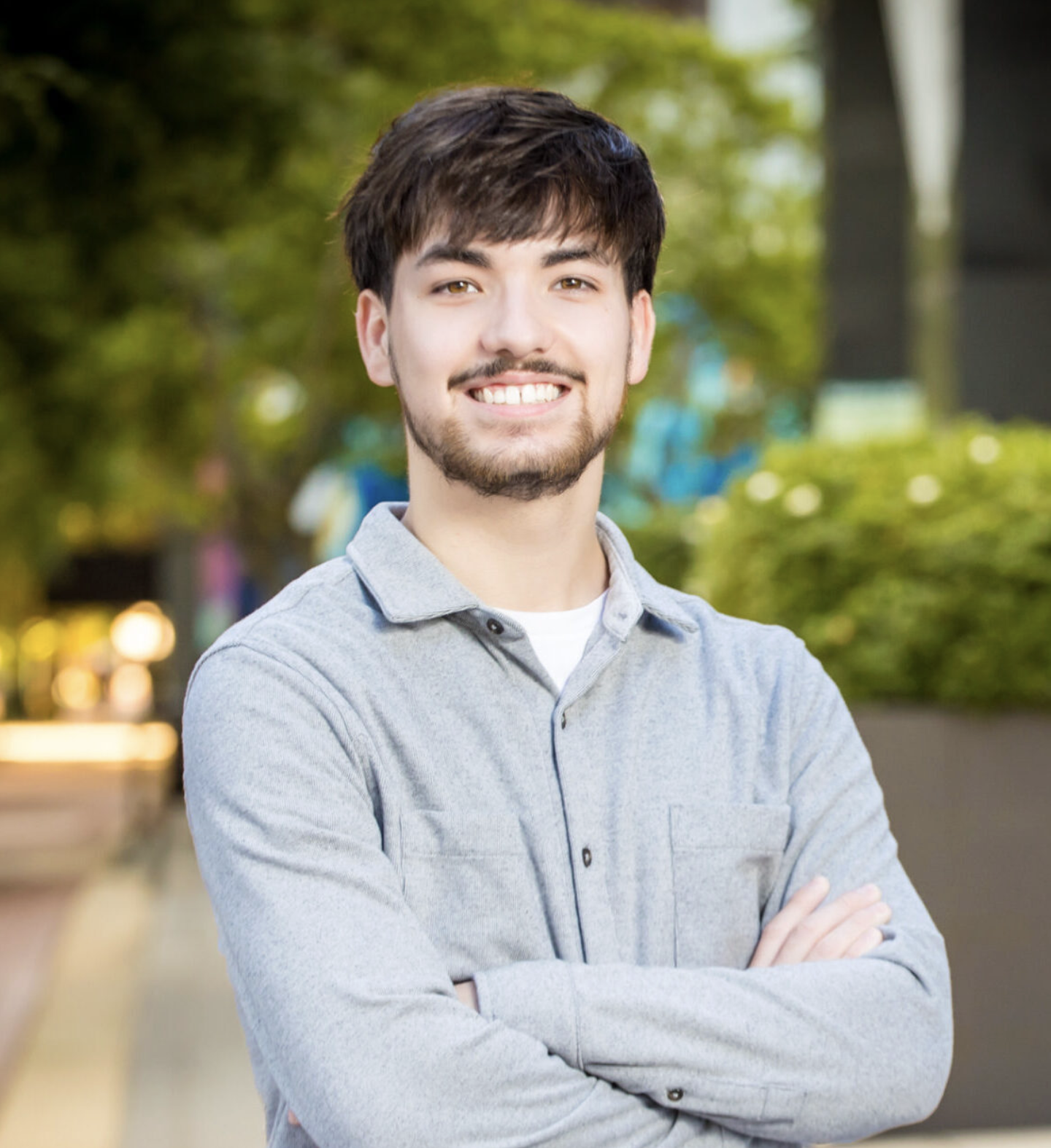 A young man (Zachary Baker) with dark hair and facial hair smiling, standing with arms crossed outdoors with blurred trees and lights in the background.