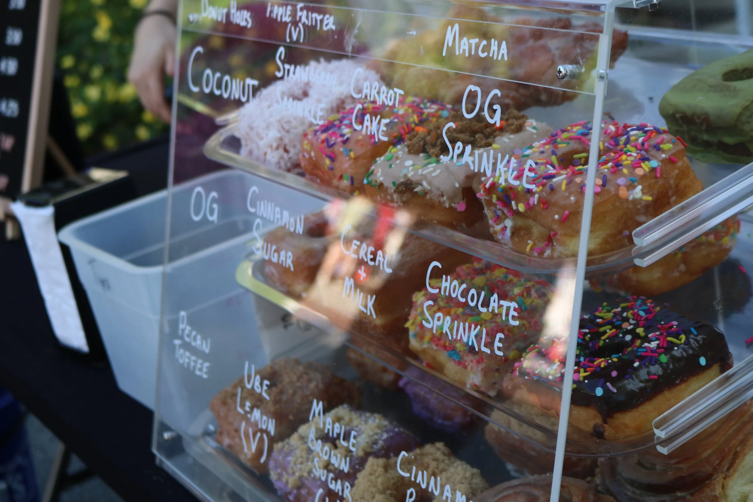 Clear display case with various donuts decorated with colorful sprinkles and icing, labeled with flavors such as 'Carrot Cake,' 'OG Sprinkles,' and 'Chocolate Sprinkle.'