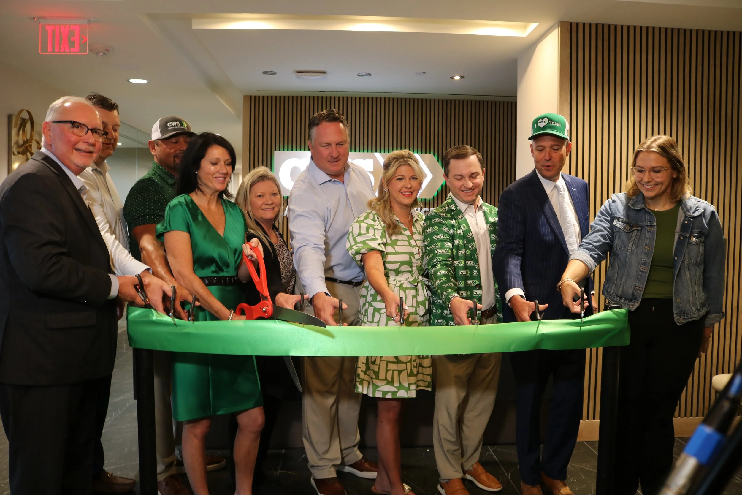 Group of people standing behind a green ribbon, participating in a ribbon-cutting event, some wearing branded hats with 'CWS' logo, in an indoor setting with wood paneling and a lit sign in the background.