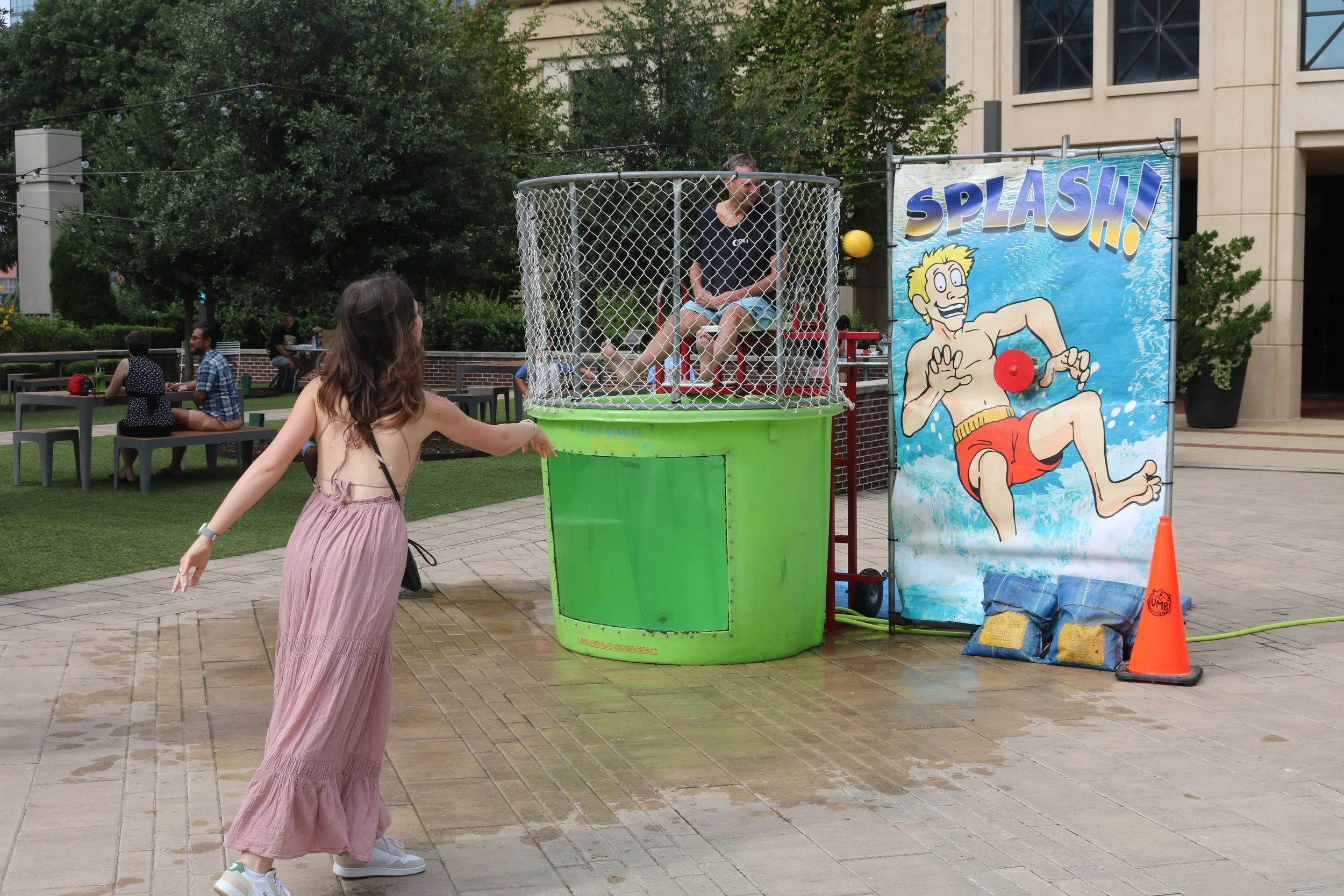 A girl in a pink dress is trying to dunk the Columbia, SC mayor in water. A colorful sign with an illustration of a cartoon boy in a swimsuit and the word "SPLASH!" is visible. People are sitting on benches in the background near trees.