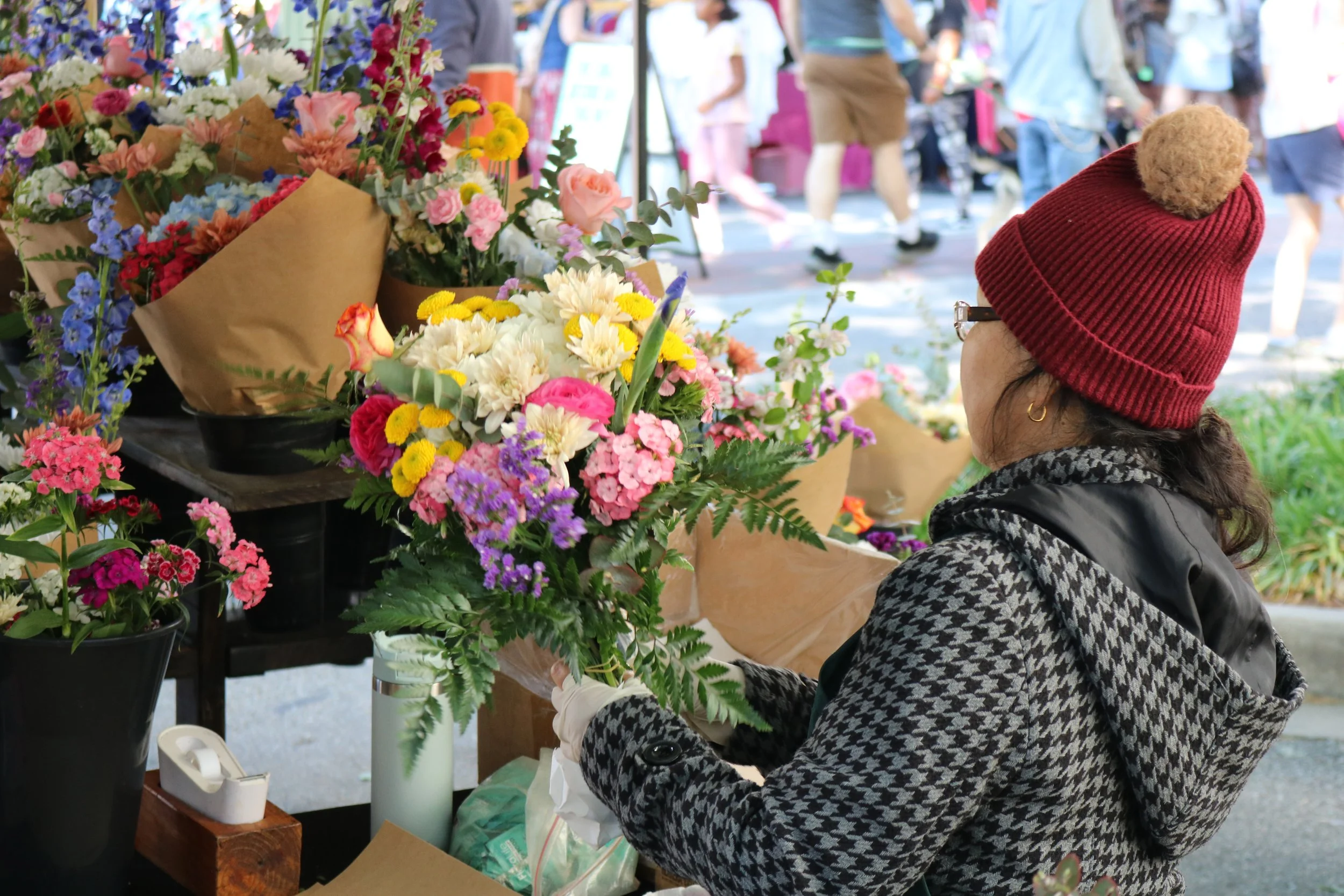 A woman with curly hair wearing glasses, a red knit hat with a pom-pom, and a black-and-white coat prepares flowers at an outdoor market. She is looking at colorful bouquets of flowers wrapped in brown paper, which are displayed on a table.