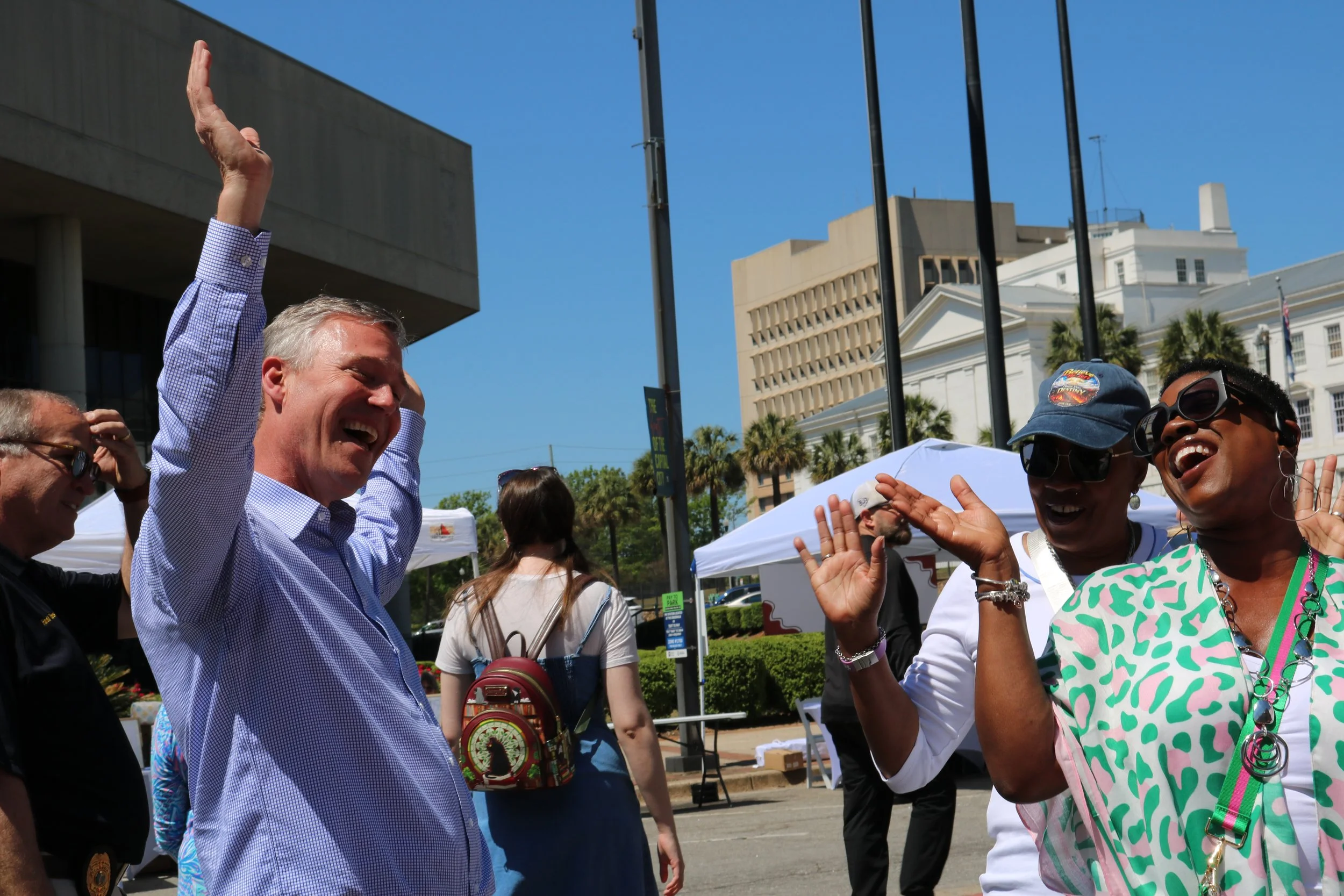 People are smiling and celebrating outdoors in a city square, with buildings and tents in the background, on a sunny day.