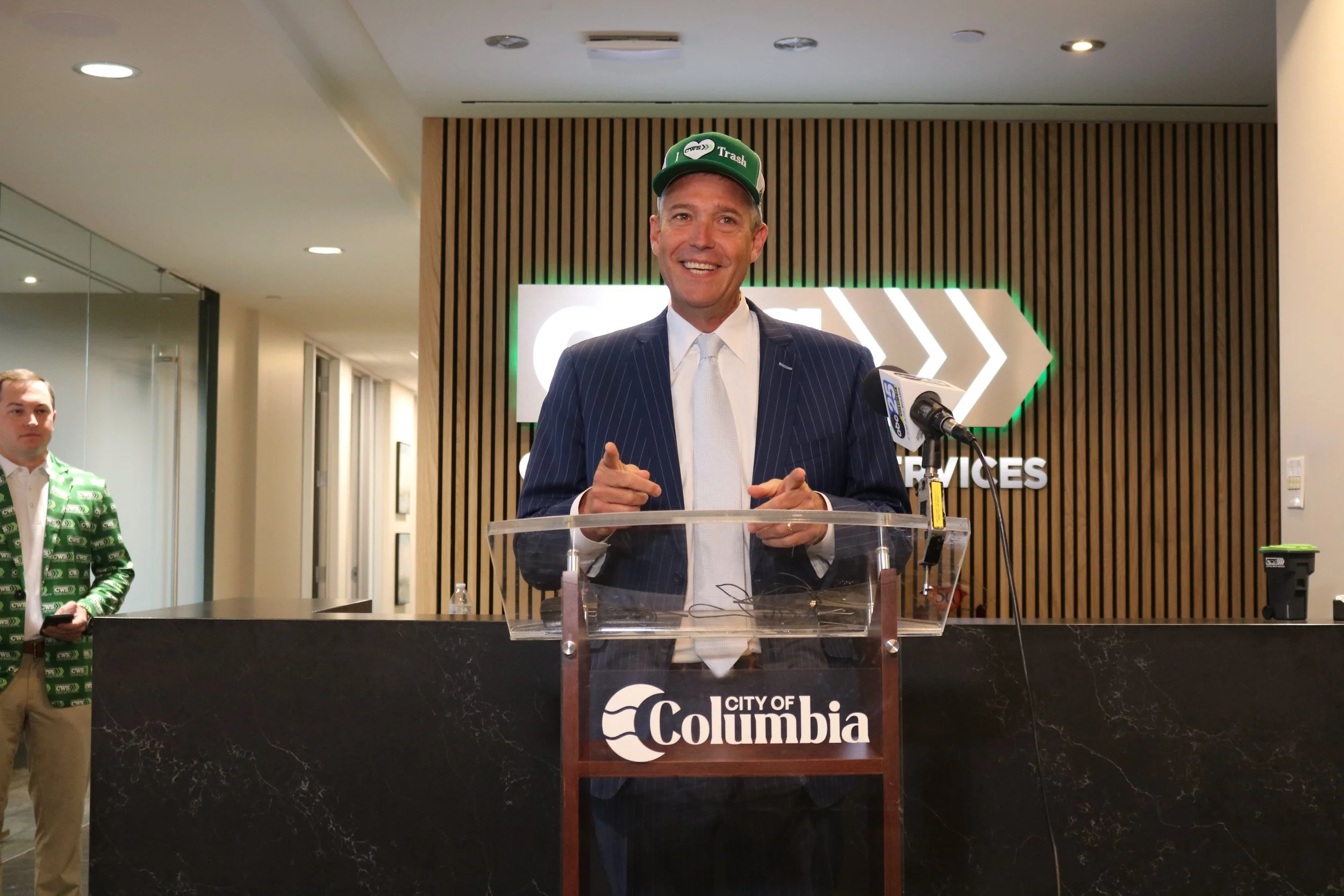 Columbia, SC mayor in dark pinstripe suit and white tie standing at a podium with a microphone, smiling and pointing. To his left is a man in a green patterned jacket. The background features a wall with wooden slats and a sign.