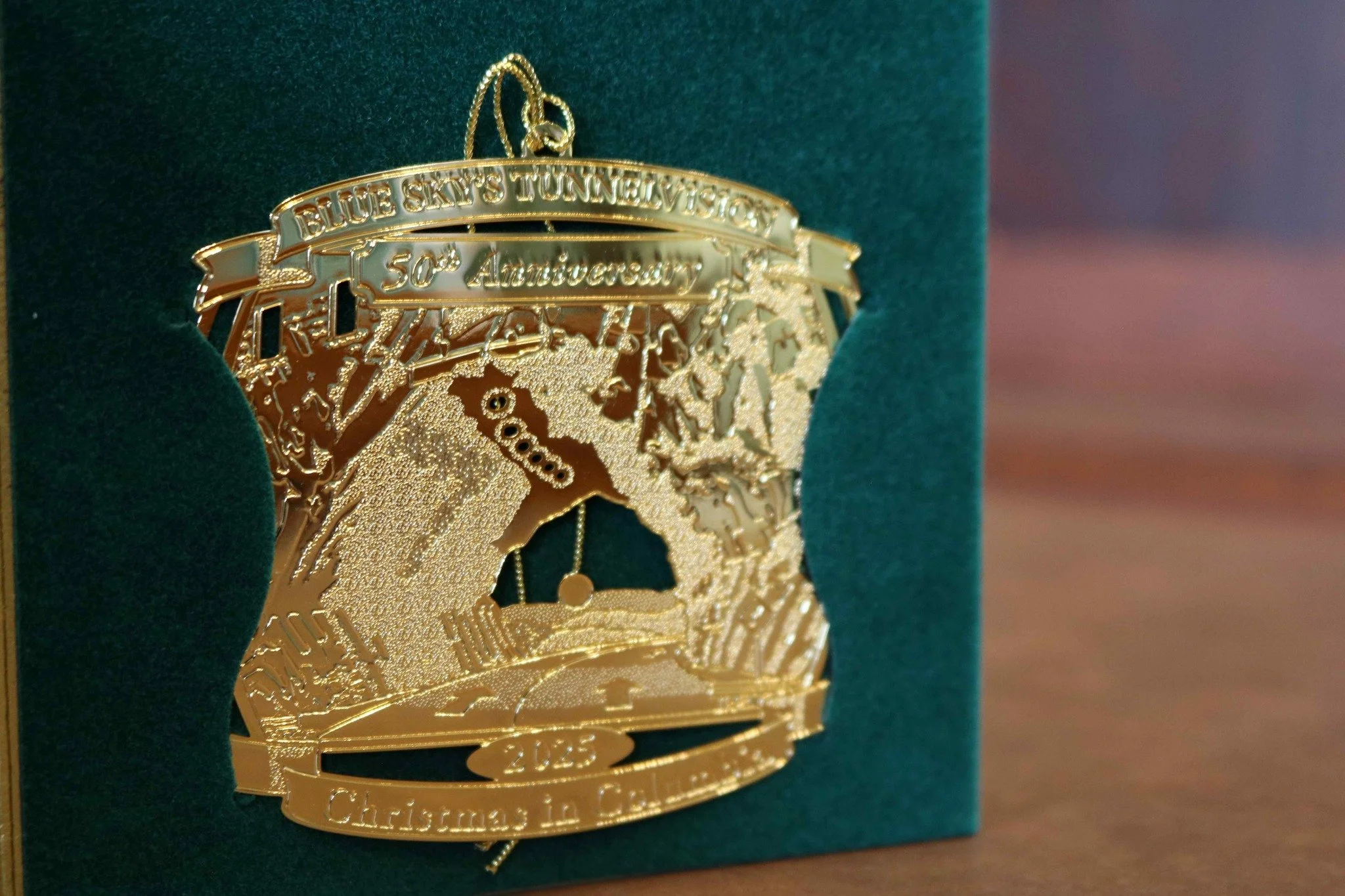 A gold-colored Christmas ornament medal with intricate designs and text that reads 'Blue Sky's Tunnels 50th Anniversary.' It is hanging on a green background.