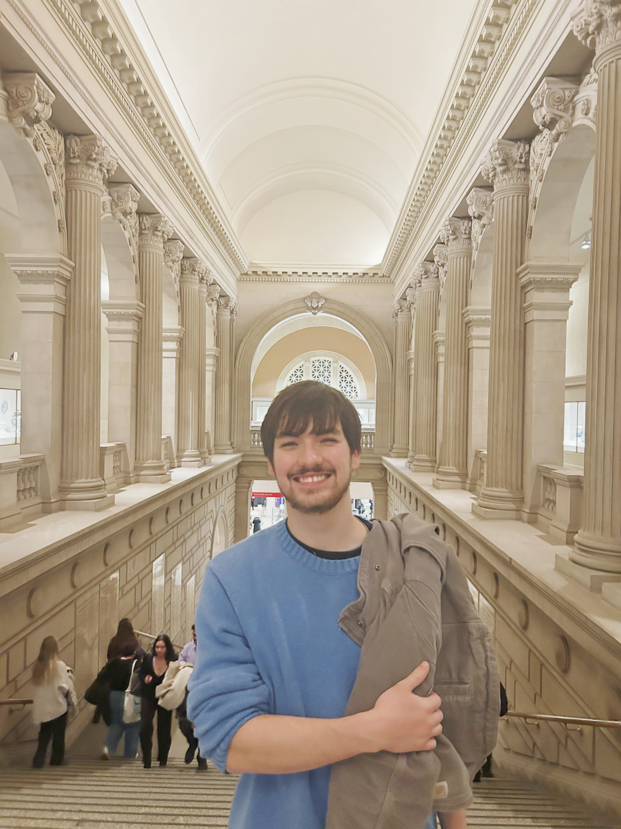 A young man (Zachary Baker) with dark hair and a beard smiling at the camera in a large ornate hall with tall columns and detailed architecture, holding a gray jacket.