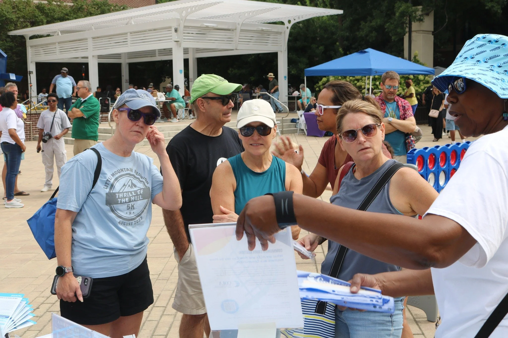 People gathered at an outdoor event, engaging with informational displays and activities, with tents in the background.