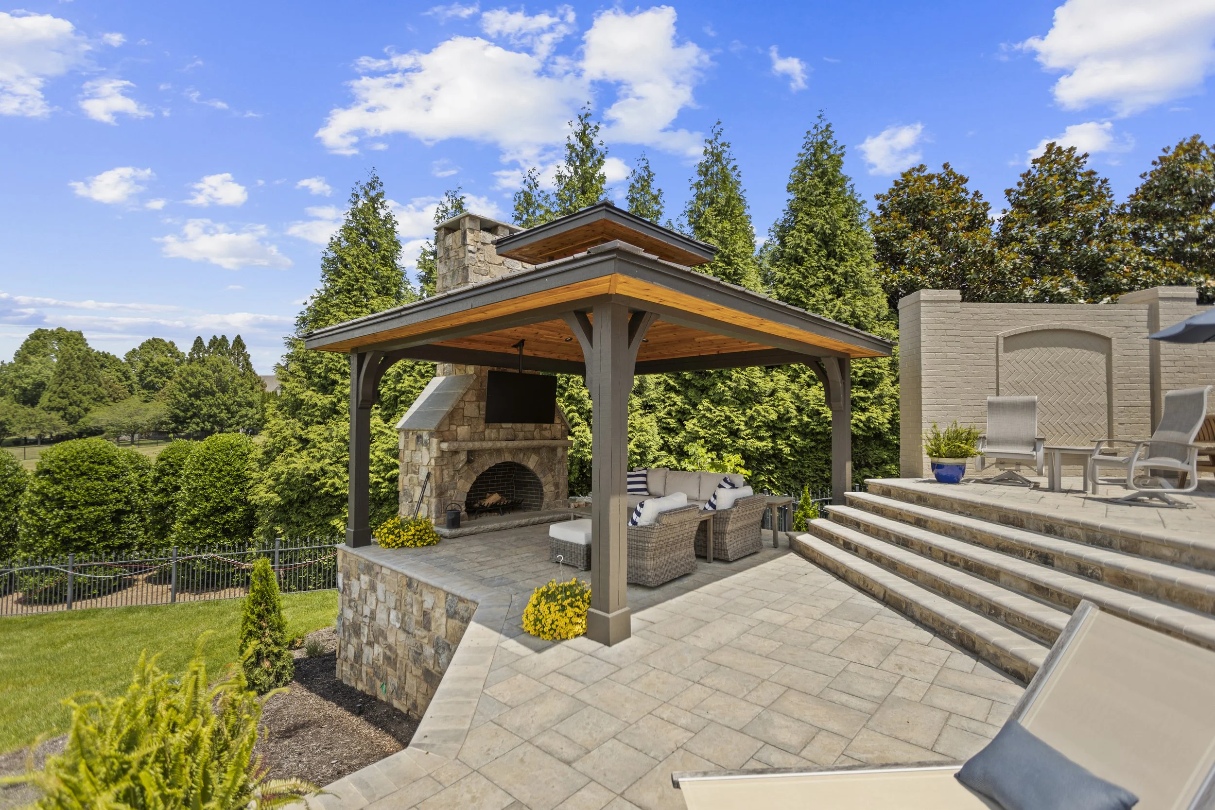 Outdoor patio with stone fireplace, seating area with chairs and cushions, potted plants, stairs, and a wooden gazebo under a blue sky with scattered clouds.