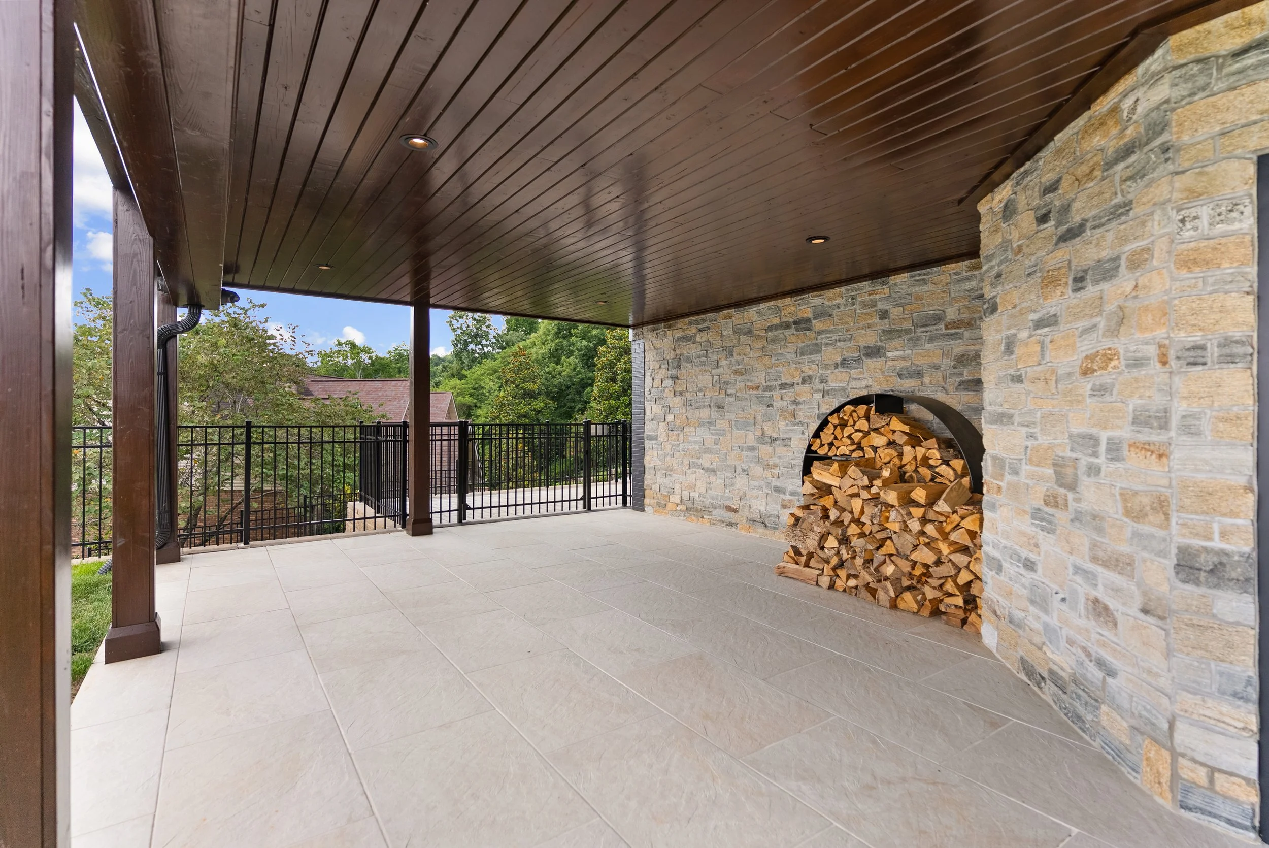 Covered patio with a wood ceiling, stone wall with firewood stacked in a rounded storage area, and black metal railing overlooking trees and houses outside.