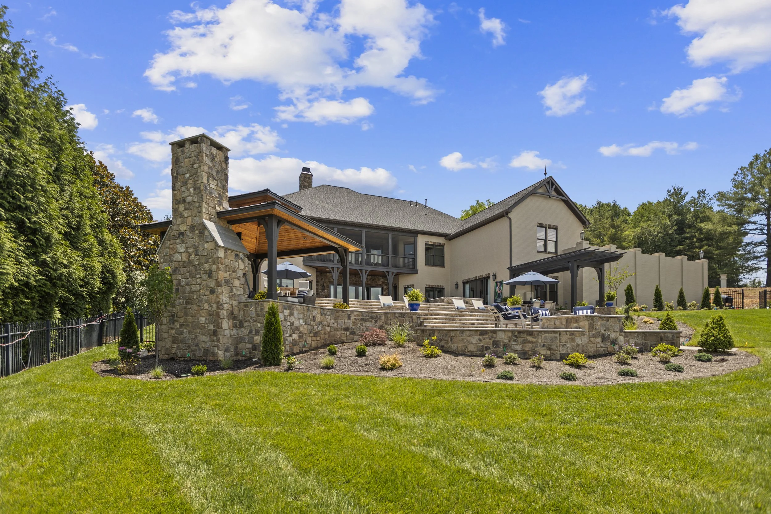 A large modern house with stone and beige exterior walls, multiple windows, a patio with outdoor furniture, umbrellas, and a well-maintained backyard with grass, shrubs, and trees under a partly cloudy blue sky.