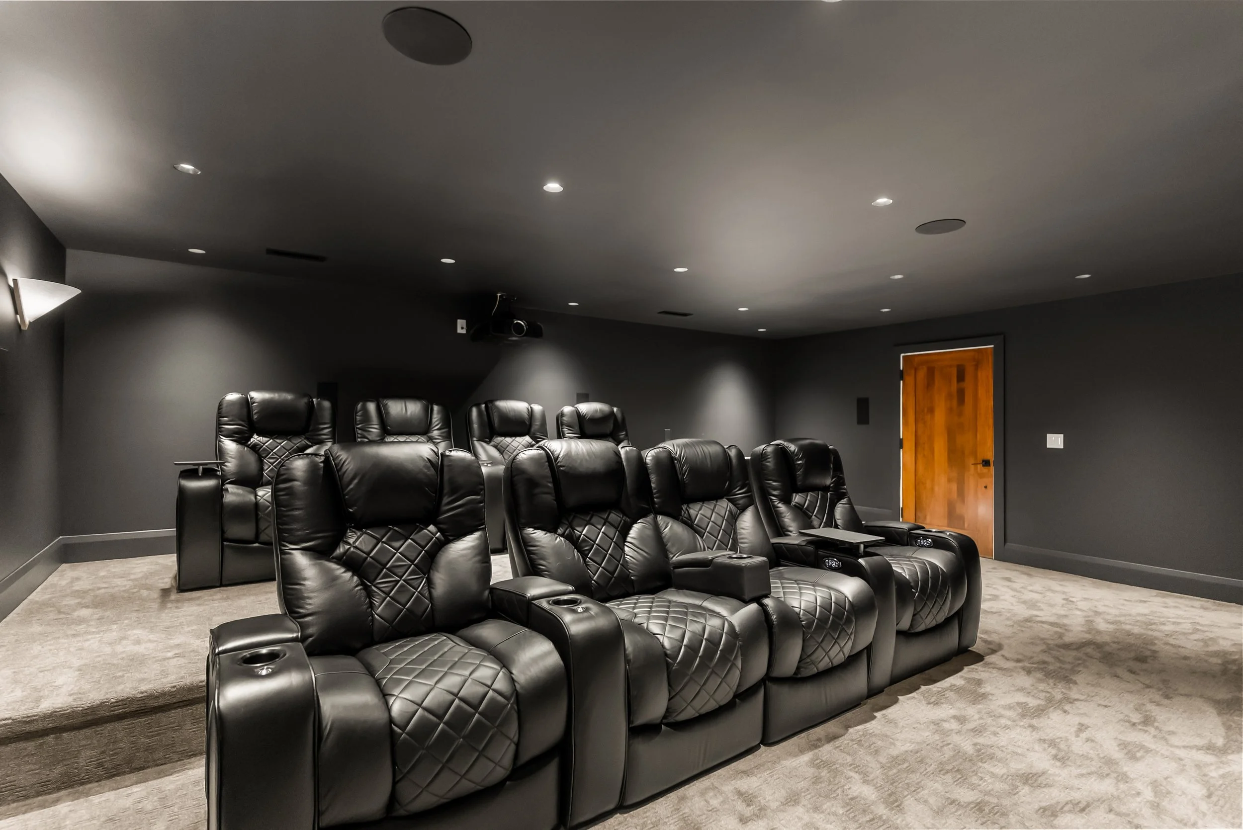 A home theater room with rows of black leather recliner chairs facing a blank wall, with a wooden door on the right side, ceiling lights, and a projector mounted on the ceiling.