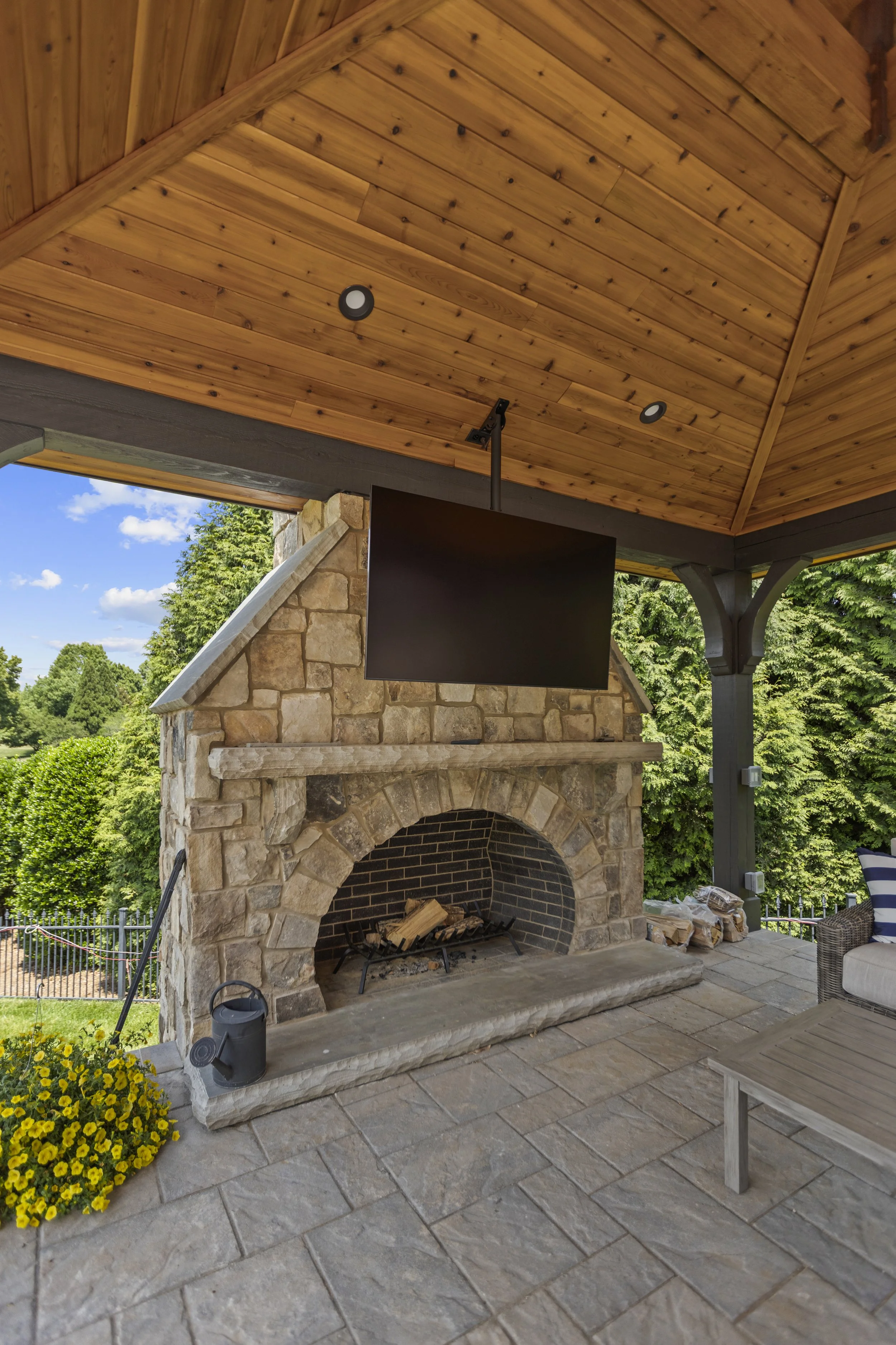 Outdoor patio with stone fireplace, hanging TV, and wooden ceiling, surrounded by greenery and yellow flowers.