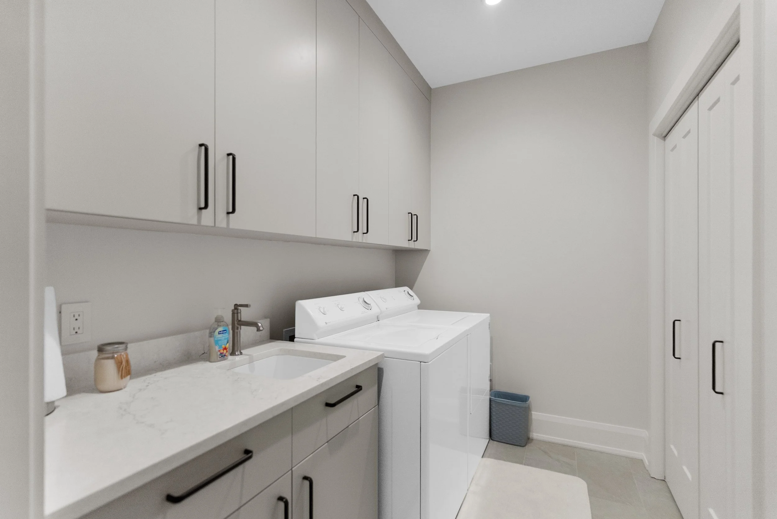 A laundry room with white cabinets, a washing machine, a countertop, a sink with a soap dispenser, a jar, and a trash bin.