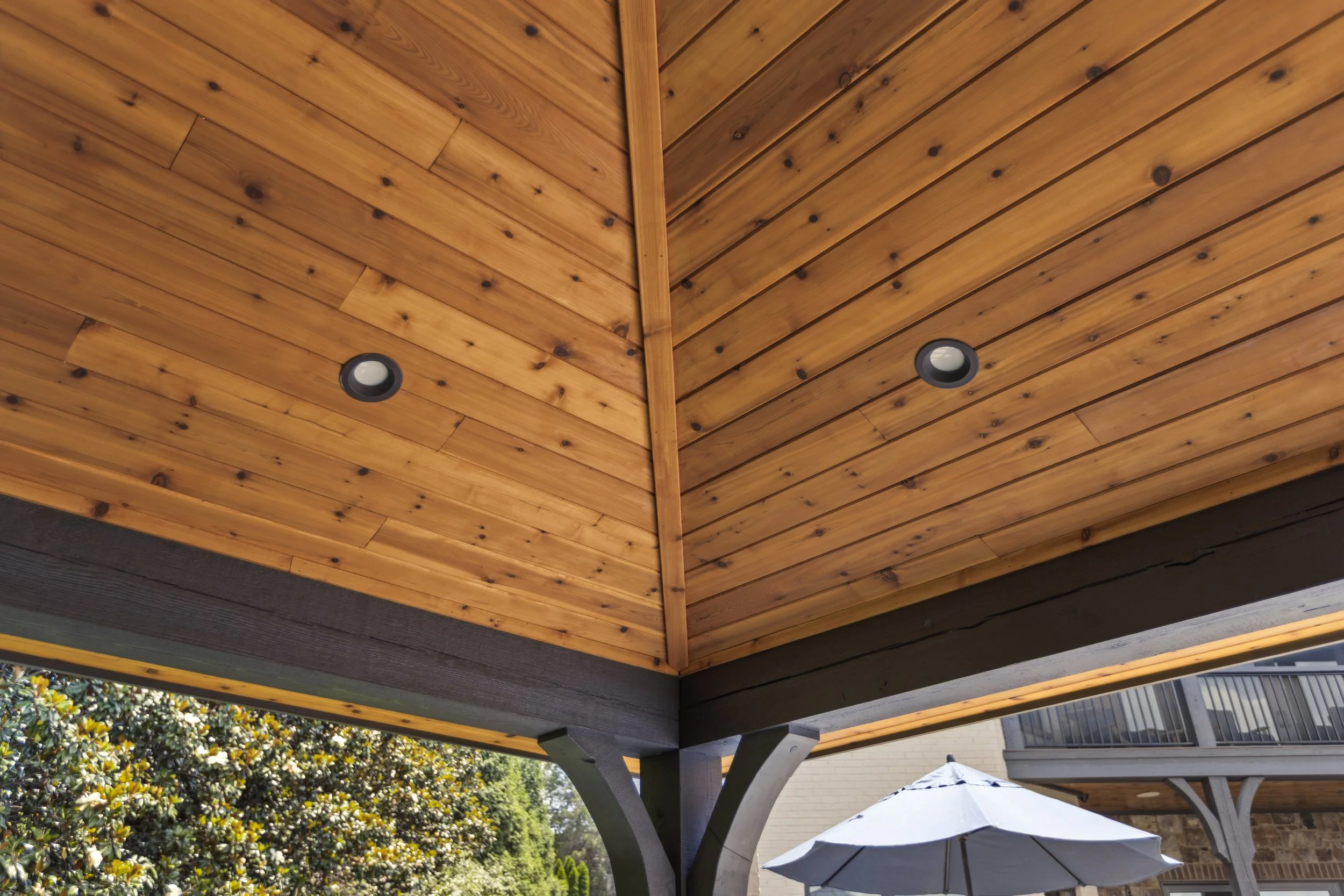 View of a wooden roof with two ceiling lights, supported by gray columns, with a patio umbrella and trees in the background.
