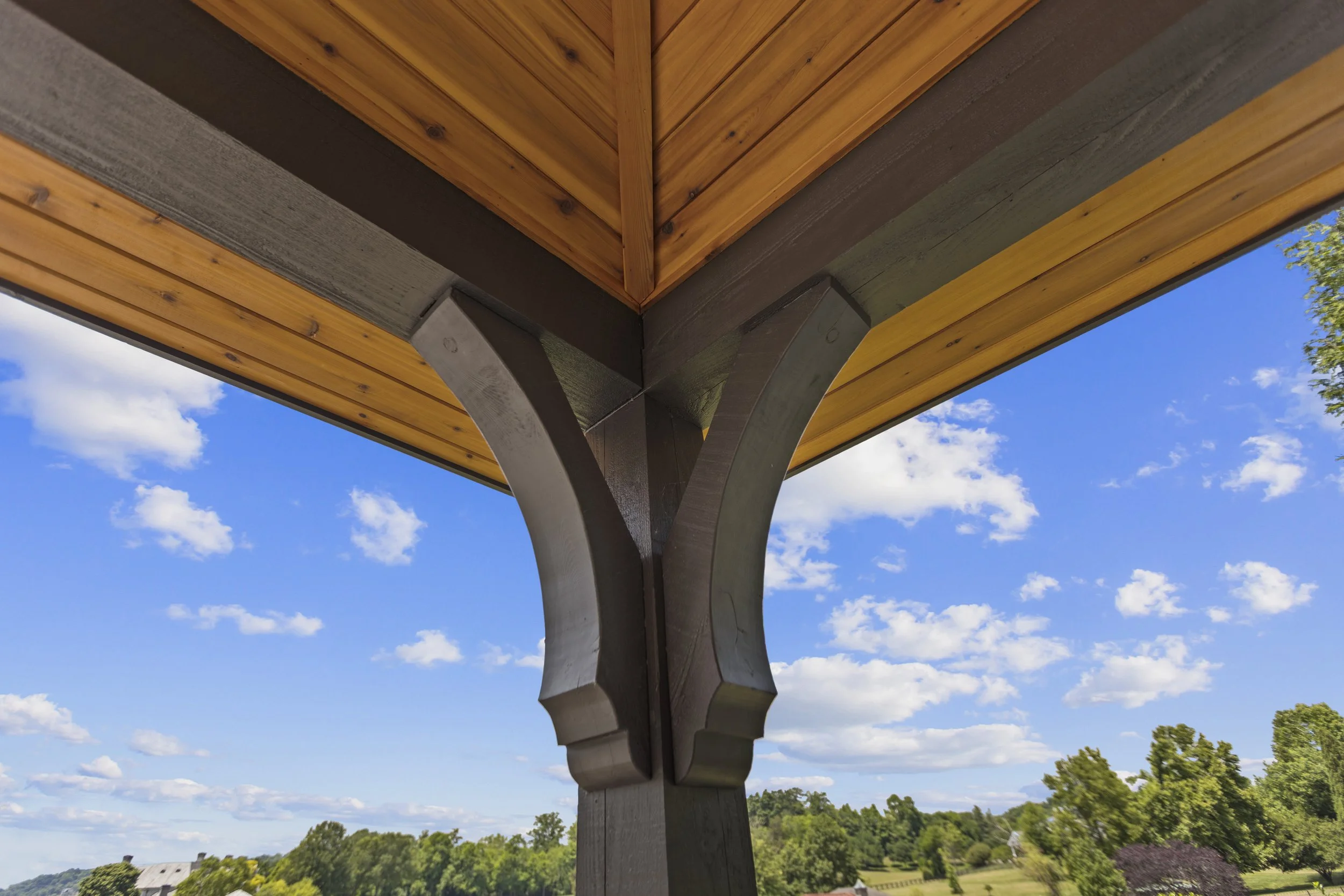 Looking up under the corner of a wooden gazebo roof, showing the wooden beams and support structures with a clear blue sky and trees in the background.