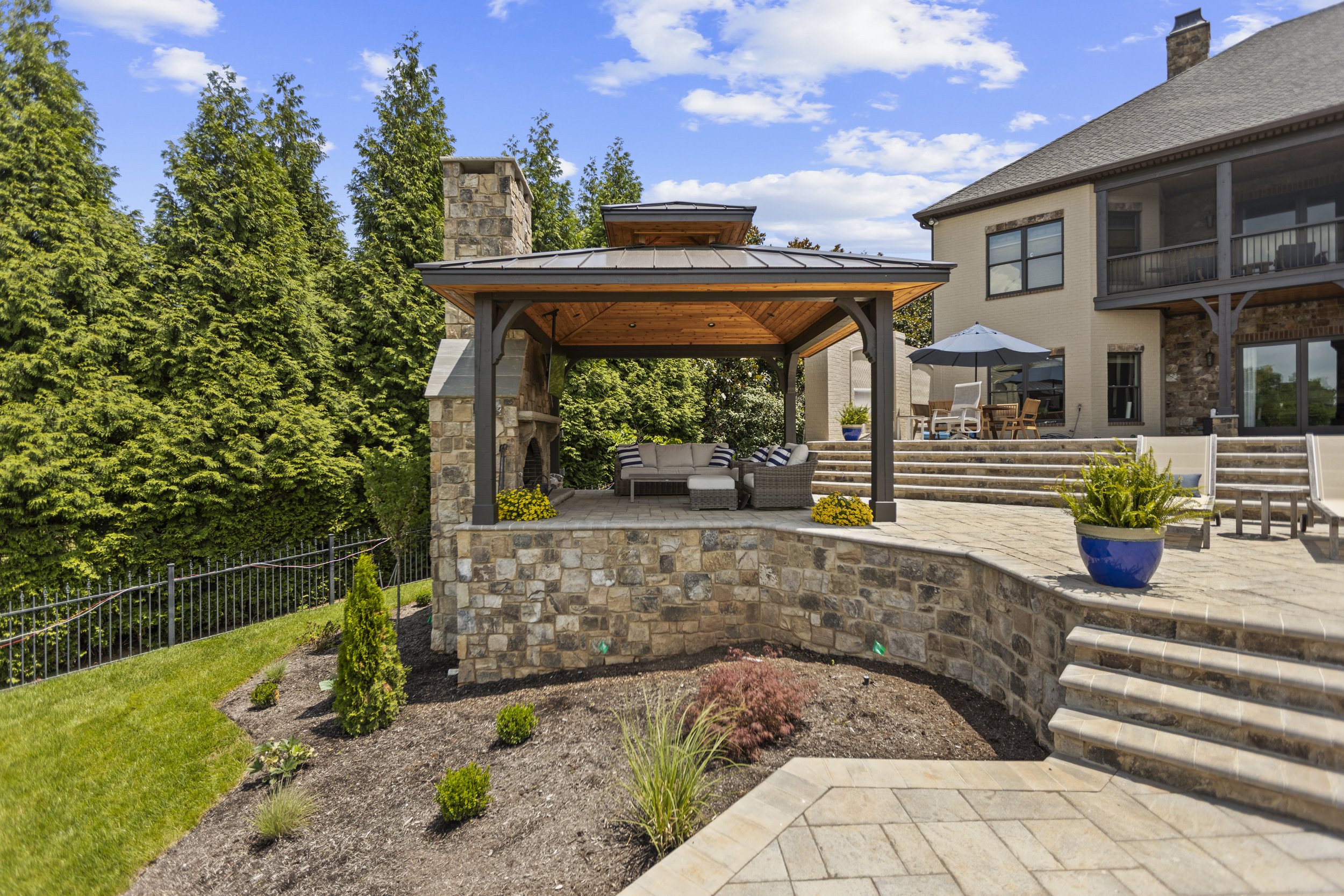Backyard patio area featuring a stone fireplace, wooden pergola, and outdoor furniture with a house in the background.