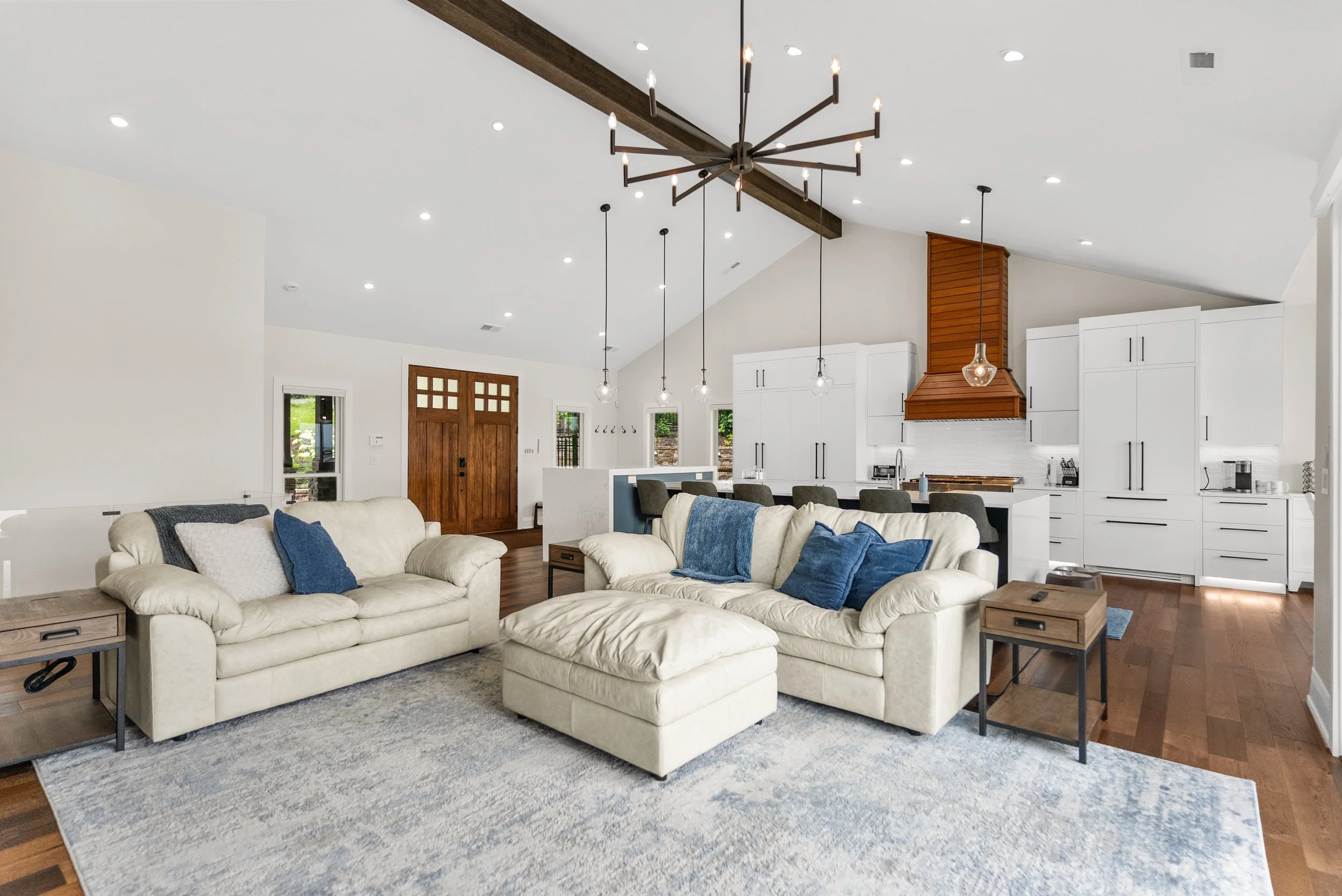 Bright open-concept living room and kitchen with a white sectional sofa, blue and beige pillows, a light blue rug, and a modern black chandelier with multiple exposed bulbs hanging from a wooden beam ceiling. The kitchen features white cabinets, a wo