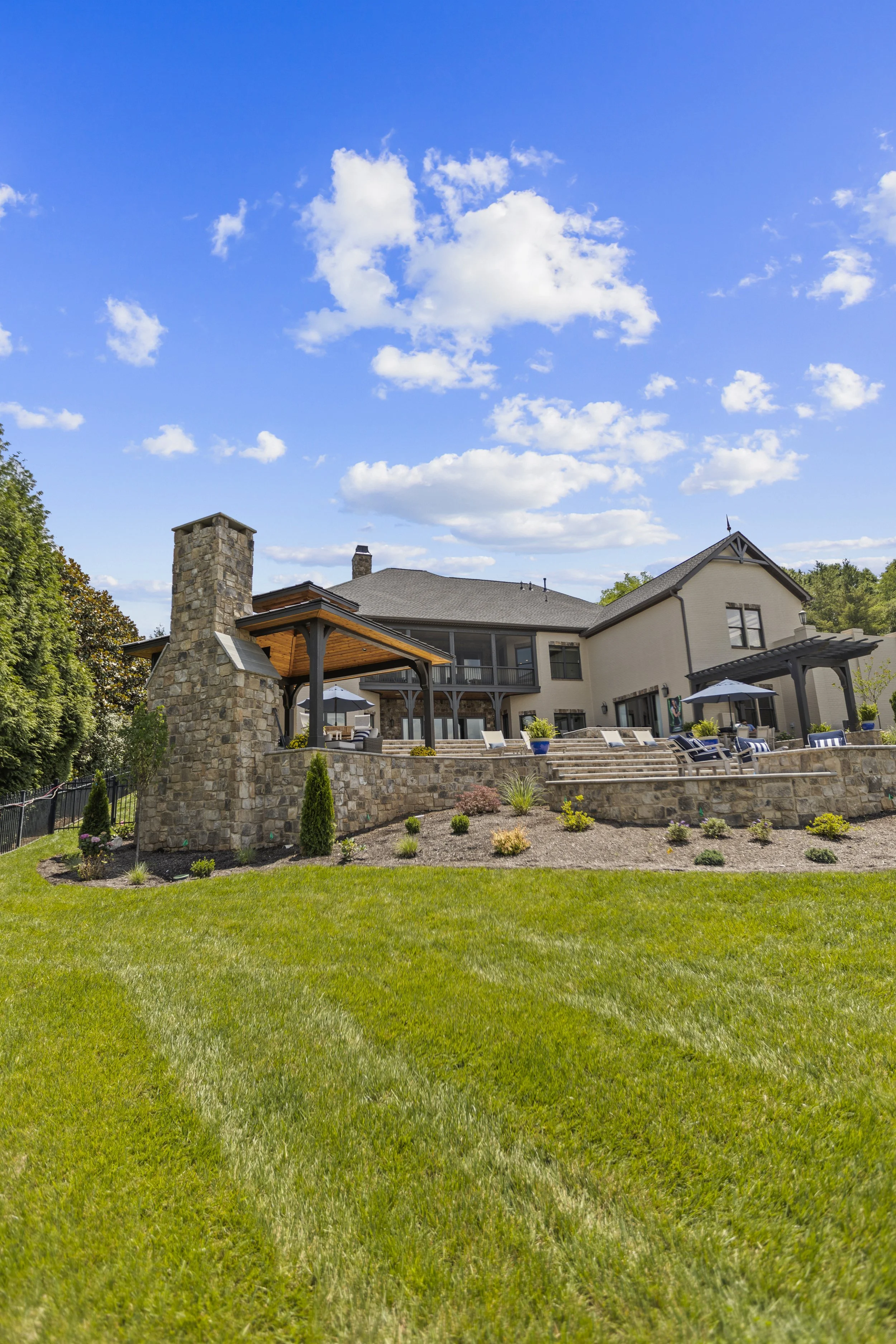 View of a backyard with a well-maintained lawn, a stone chimney, a house with a balcony, outdoor seating area with umbrellas, and a blue sky with scattered clouds.
