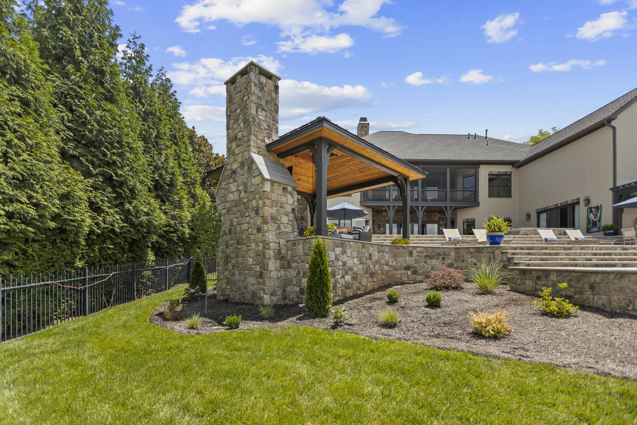 A backyard with a stone patio, landscaped garden, and a large beige house with outdoor seating and a balcony. There is a tall stone chimney, lush green trees, and a blue sky with clouds.