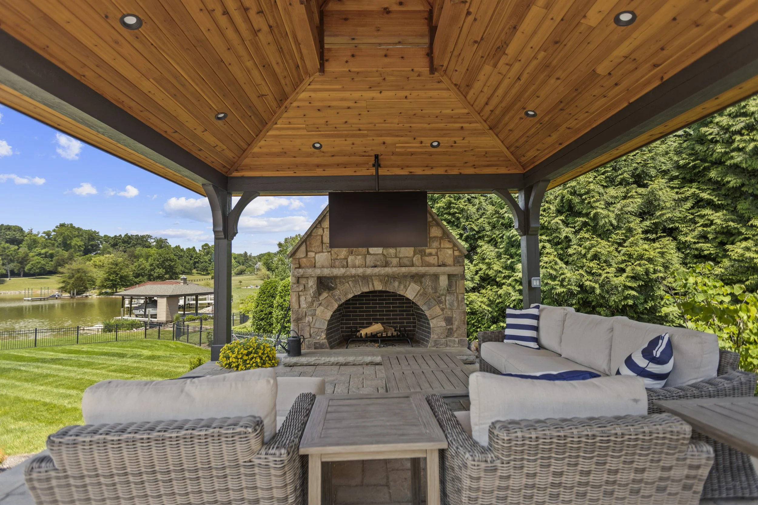 Covered outdoor patio with a ceiling of wood panels, built-in ceiling lights, a stone fireplace with wood logs, a white cushioned outdoor sofa with striped pillows, and surrounding greenery overlooking a lake.