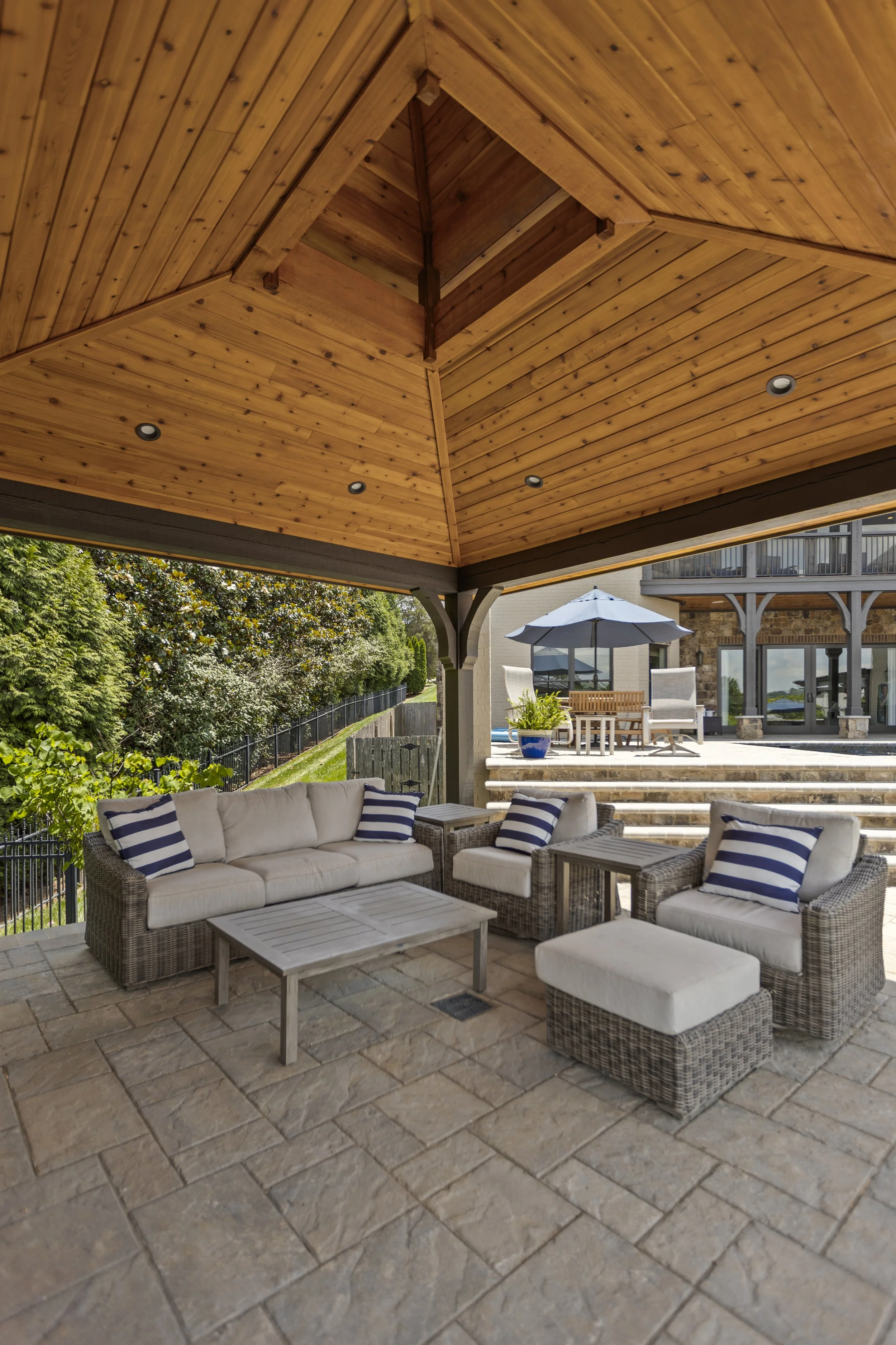 Outdoor patio with wicker seating and cushions, a wooden ceiling with recessed lighting, and a view of a garden and house with a porch and umbrella in the background.