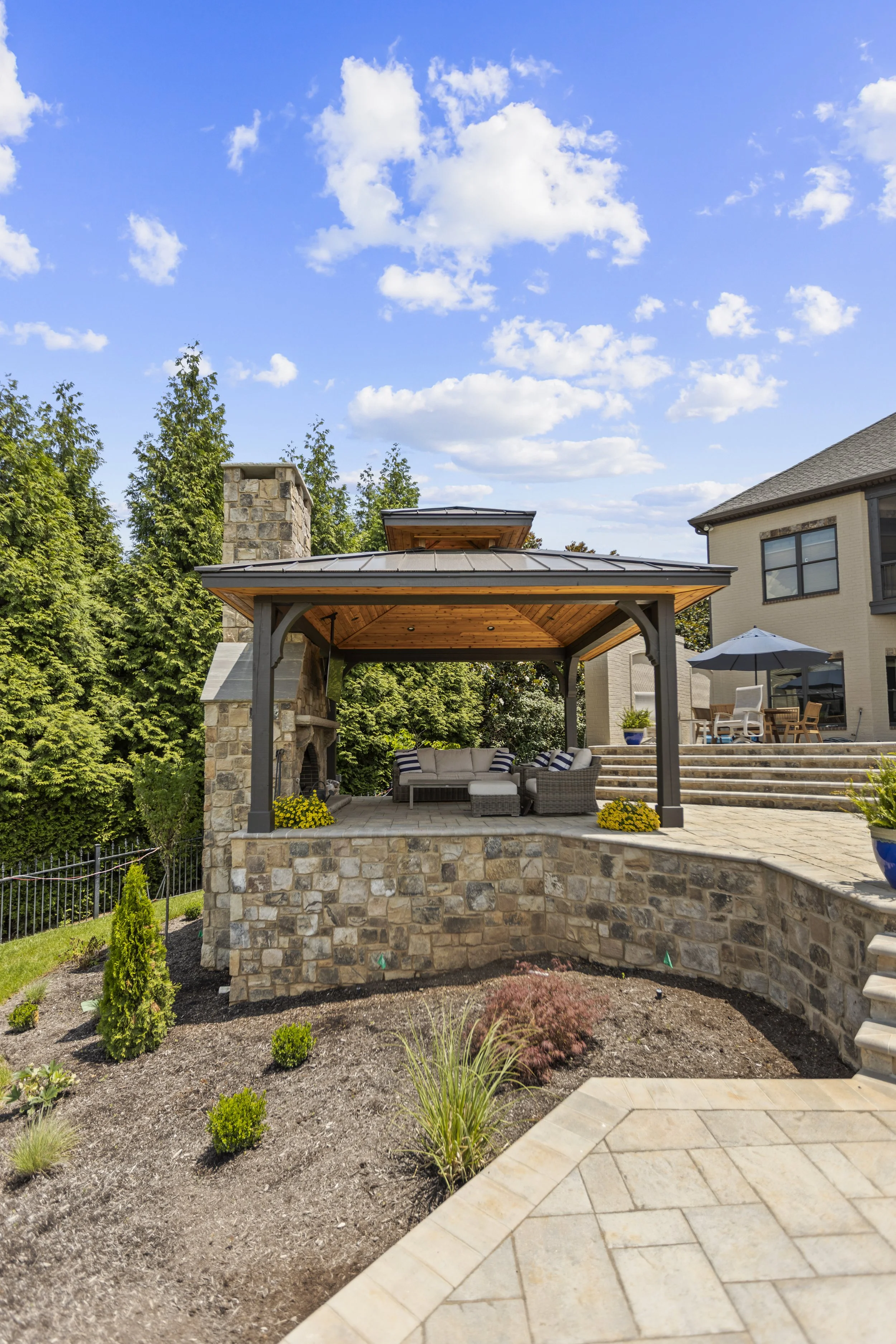 A backyard patio area with a stone fireplace and built-in outdoor seating under a wooden roof structure. There are potted plants and a well-maintained garden with small shrubs and flowers. In the background, a house with large windows and outdoor fur