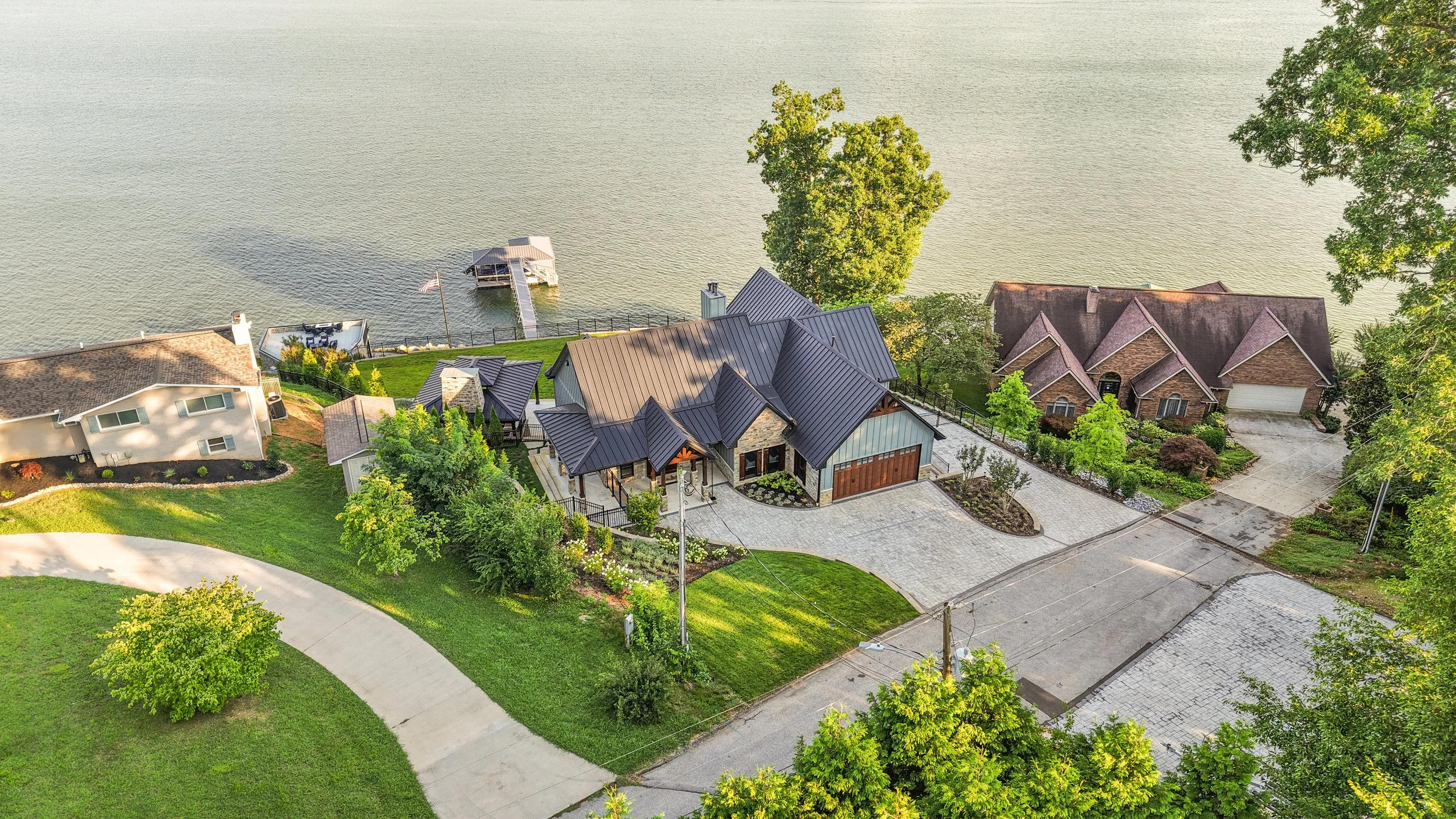 Aerial view of lakeside residential houses with green lawns, trees, and a dock extending into the water.