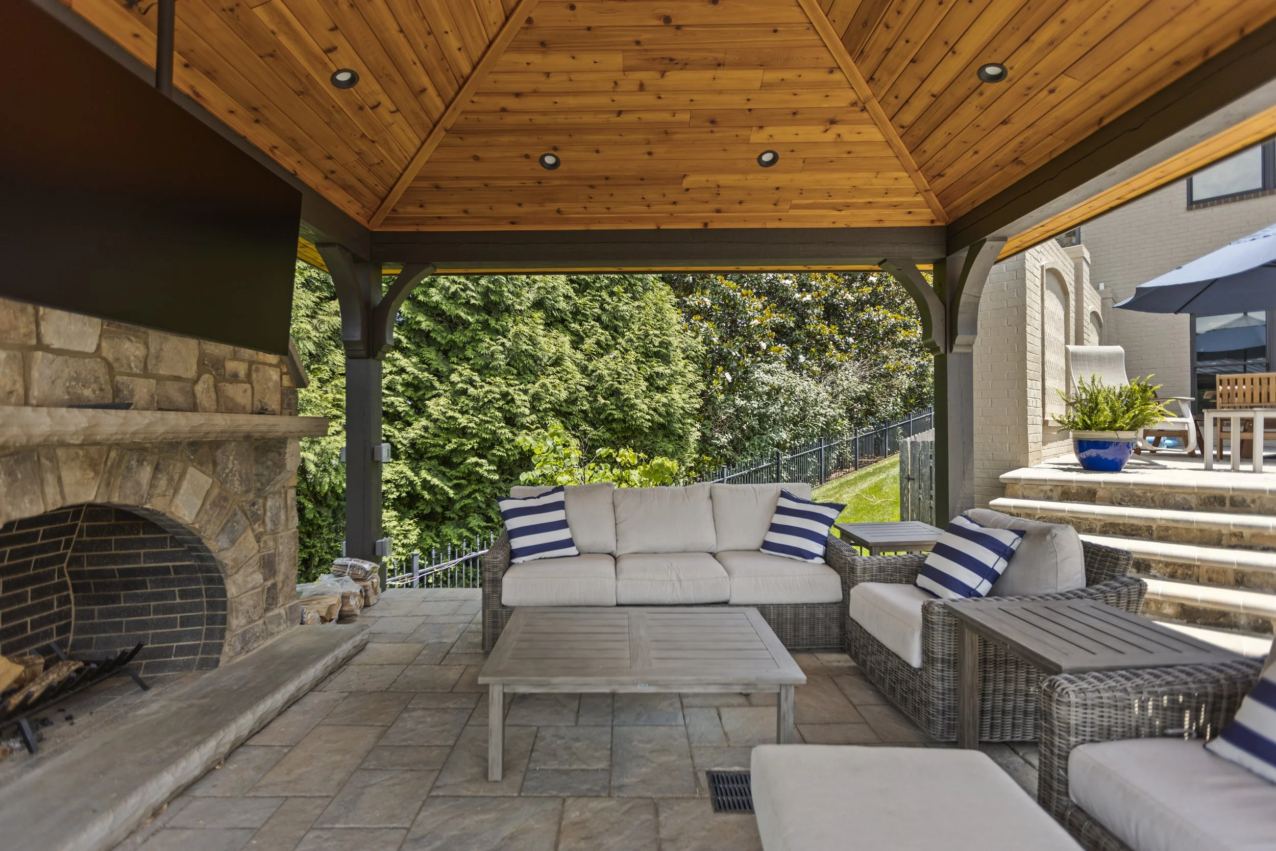 Covered outdoor patio with wicker furniture, a stone fireplace, and a wooden ceiling, surrounded by trees and a residential yard with stairs and an umbrella.