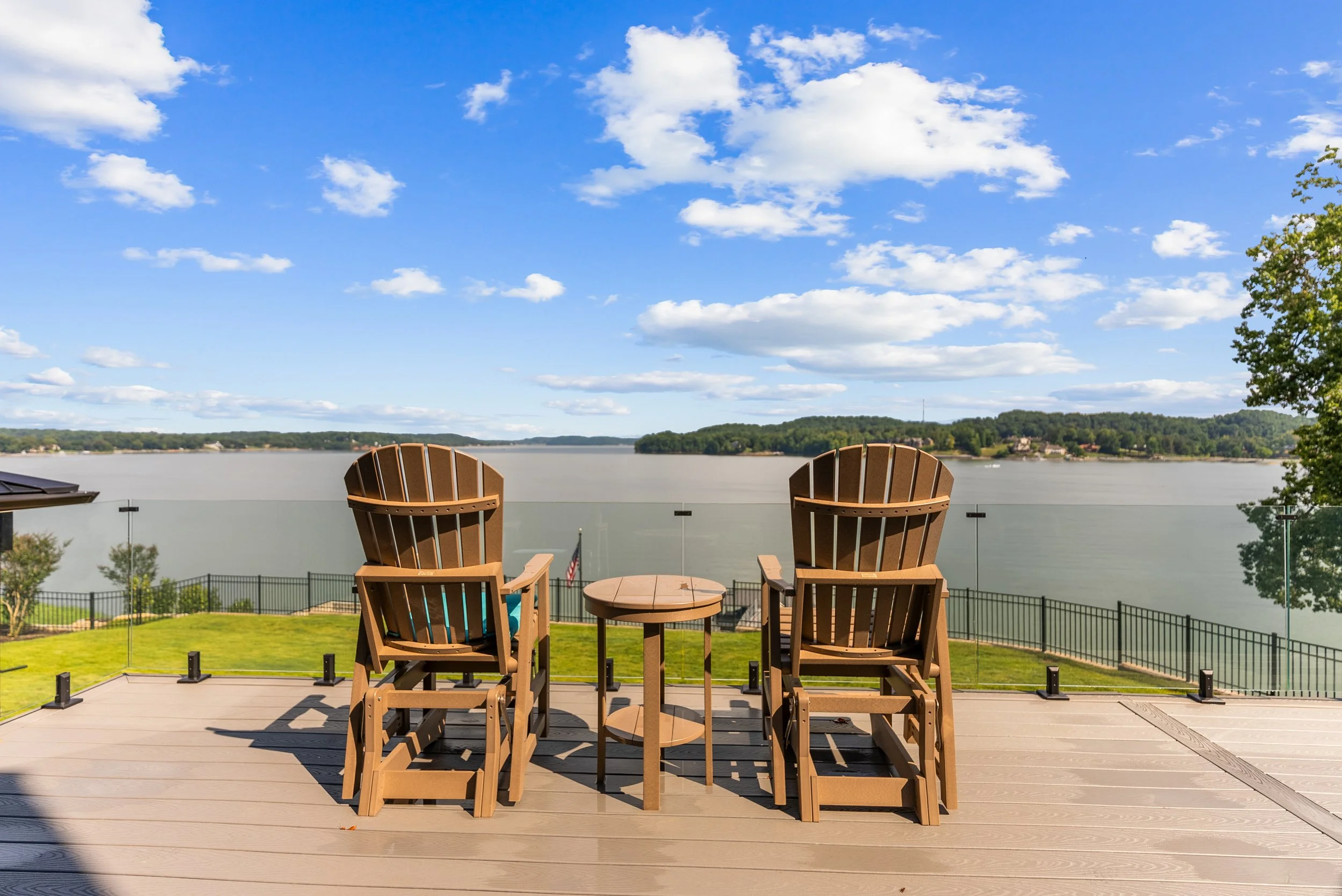 Two wooden Adirondack chairs and a small round table on a deck overlooking a river, with green hills and trees on the far bank and a partly cloudy sky overhead.