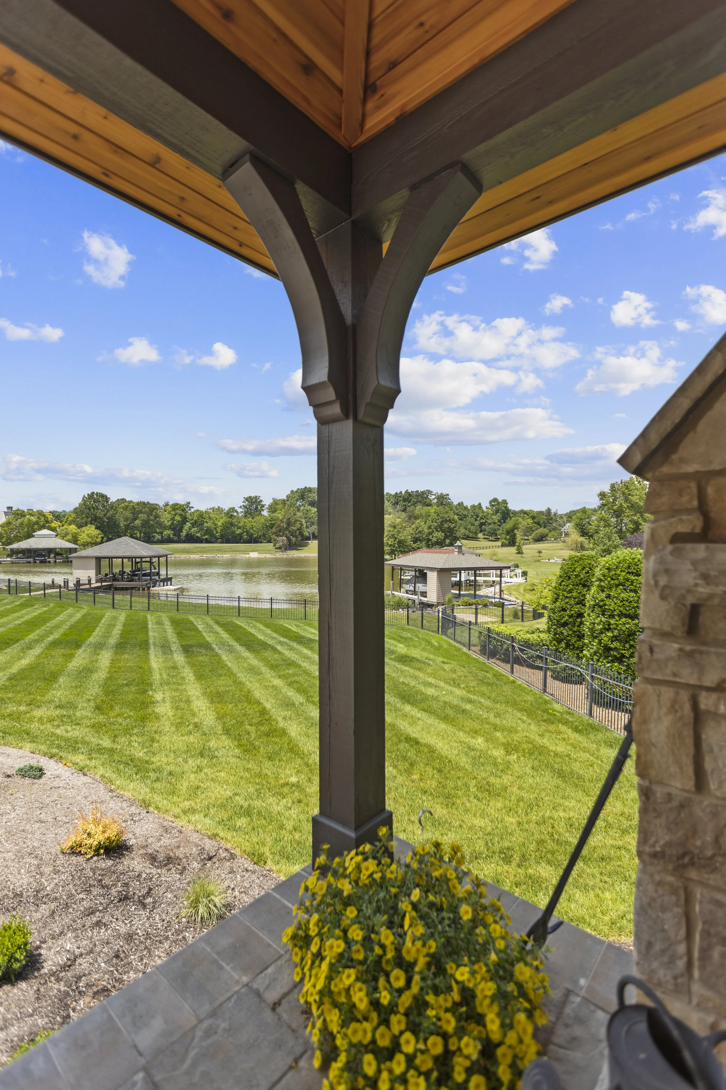 View from under a building porch showing a landscape with a river, green trees, and a gazebo in the background. The porch has a flower pot with yellow flowers and a stone wall on the right.