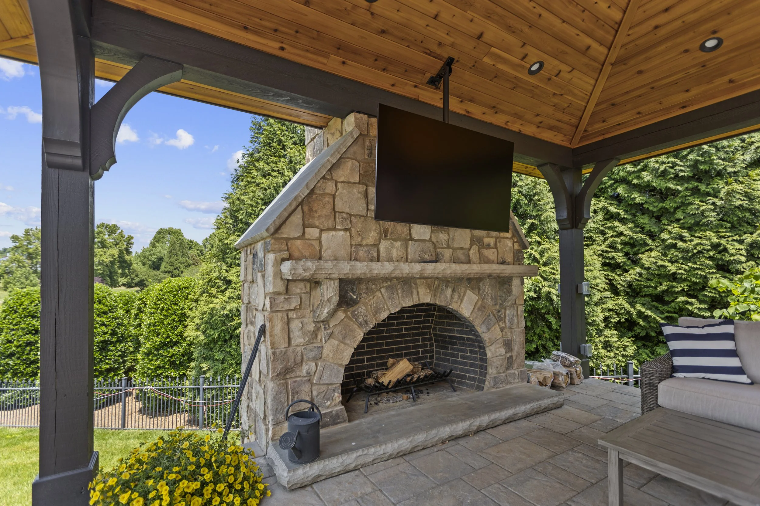 Outdoor covered patio with stone fireplace, wooden ceiling, and outdoor furniture, surrounded by green trees and blue sky.