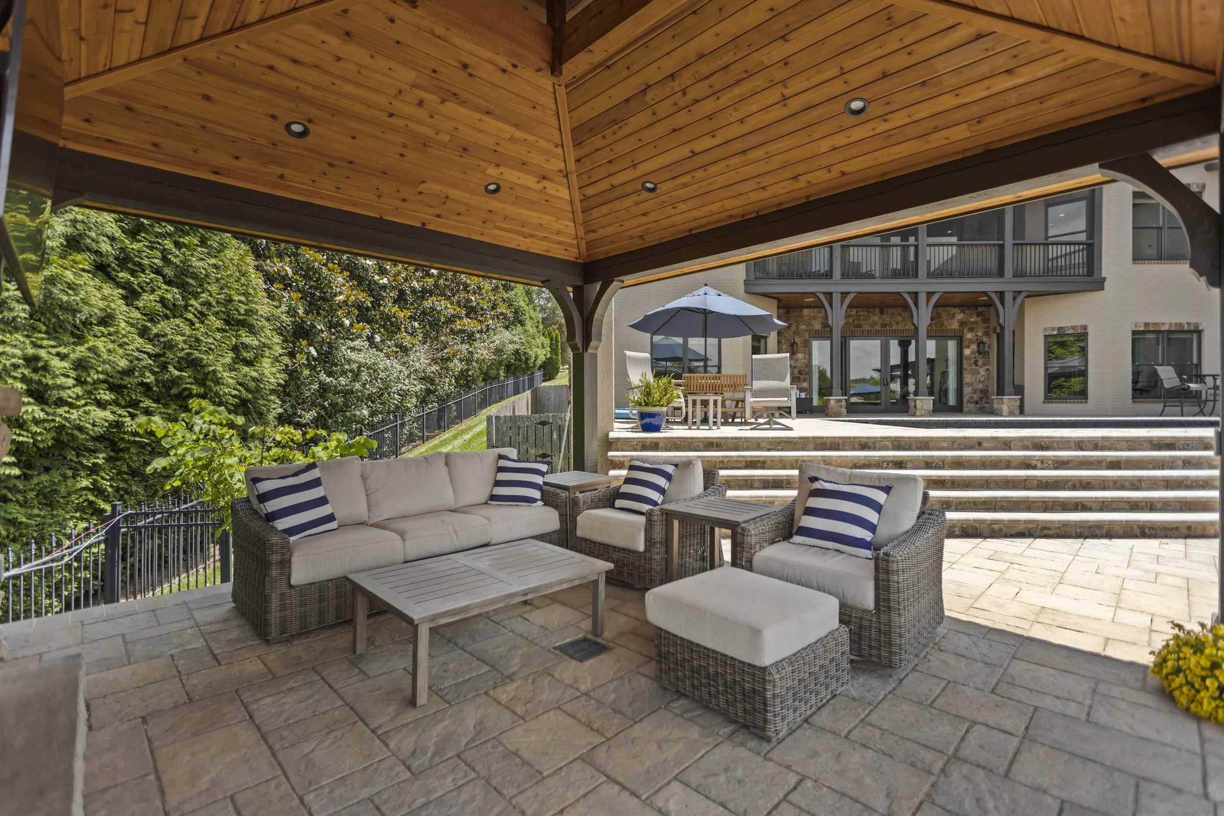Outdoor patio area with wicker furniture, striped pillows, a stone patio, wooden ceiling, and a view of stairs, a house, and greenery.