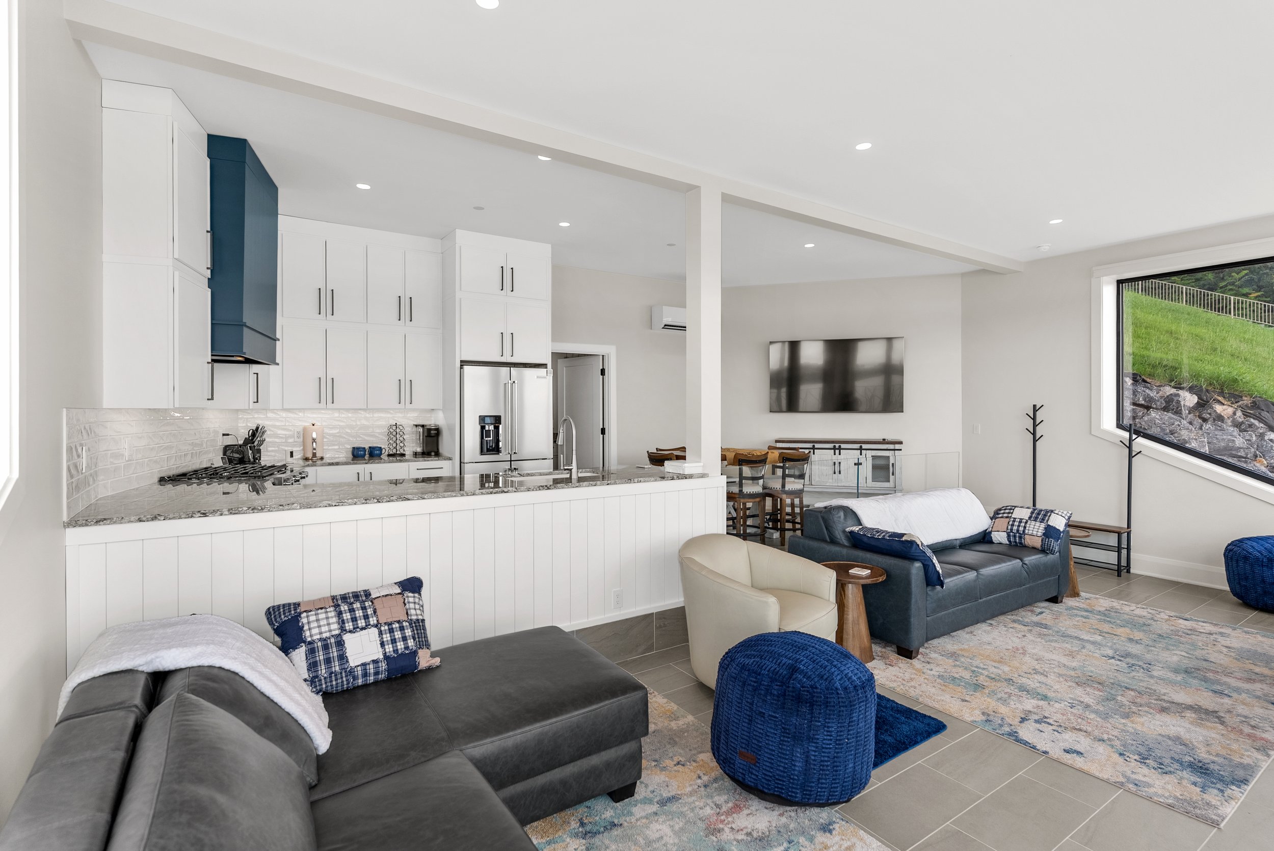 Open living room and kitchen area with modern furniture, large window showing a green landscape, white cabinets, and a wall-mounted TV.
