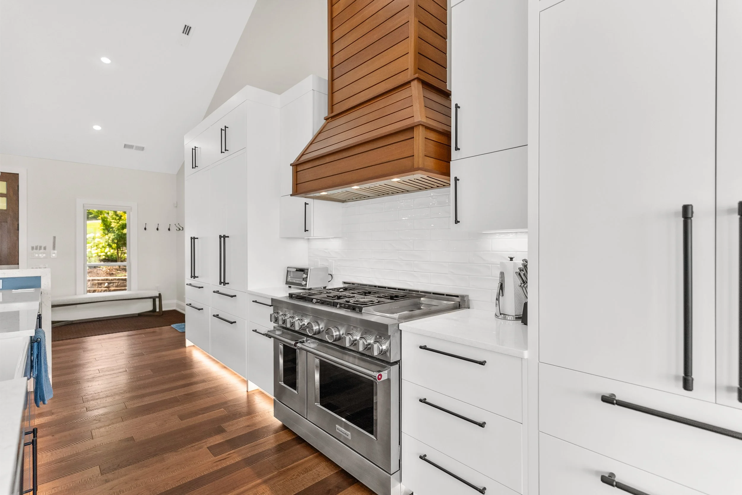 Modern kitchen with white cabinets, black handles, a stainless steel gas stove, and wooden flooring. There is a wooden range hood, a small toaster, and a coffee maker on the white countertop. A window shows greenery outside.