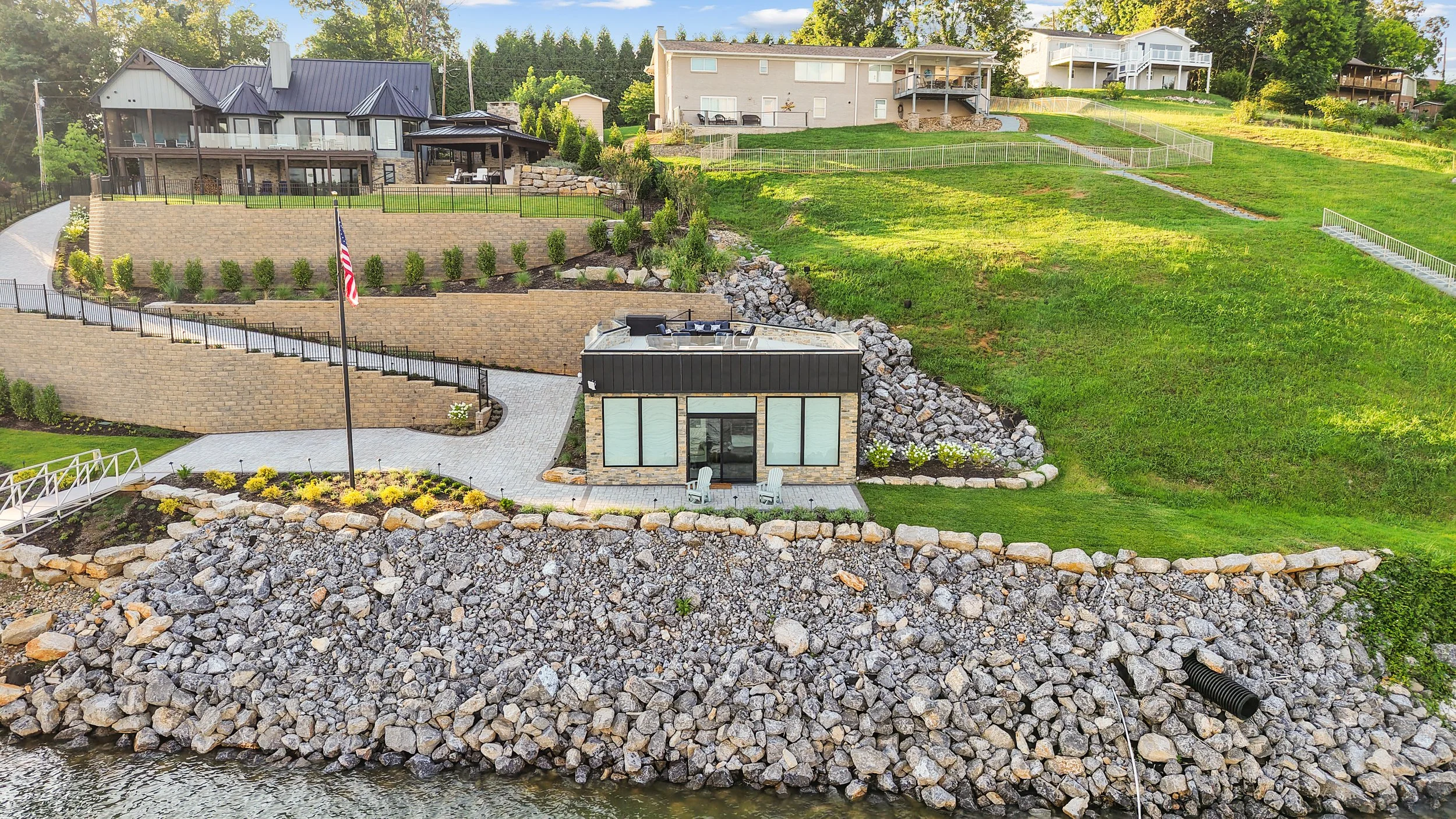 Residential hillside with multiple modern houses, manicured lawns, and landscaping, adjacent to a rocky shoreline by a body of water.