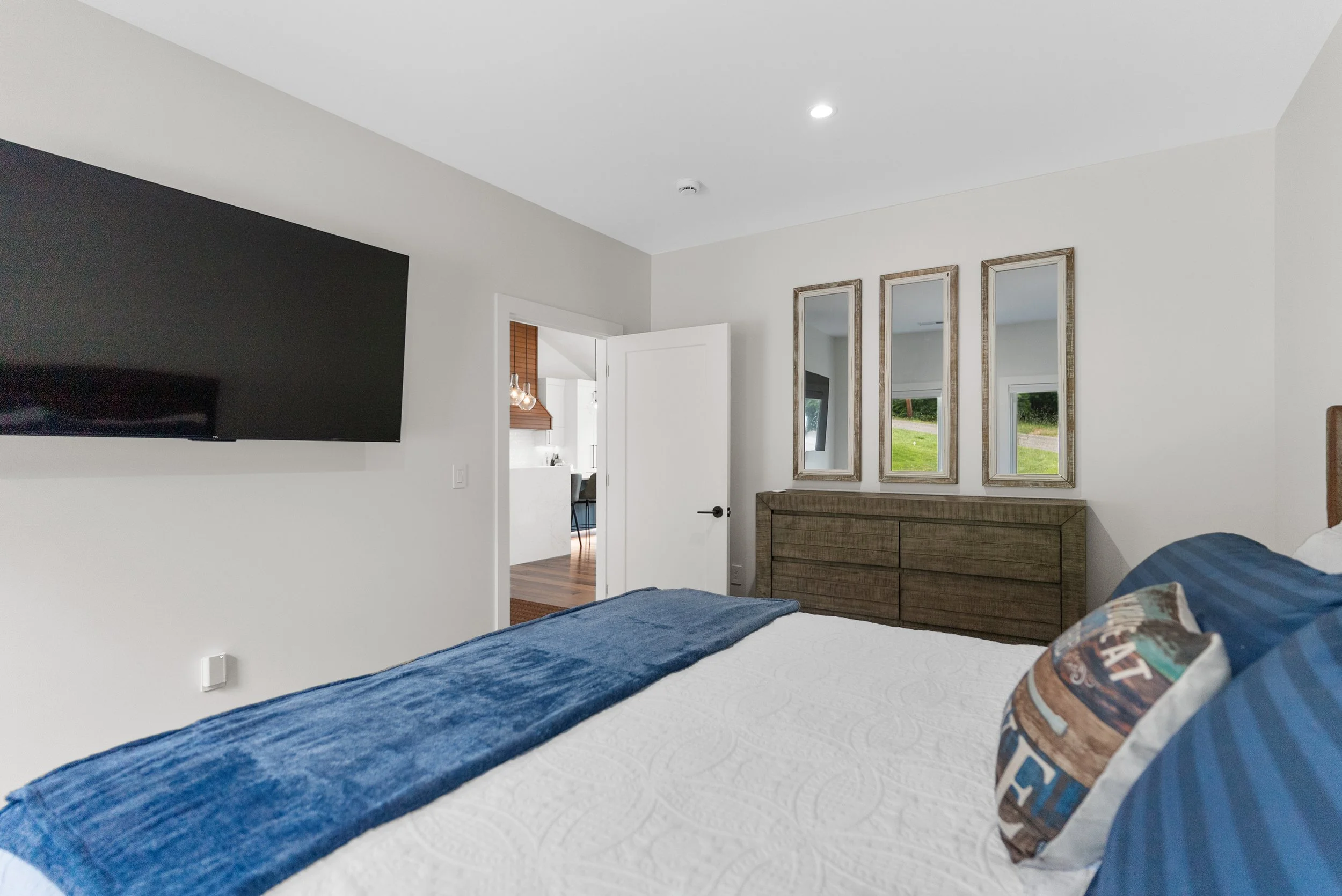 Bedroom with a white bedspread, blue quilt, patterned pillow, dresser with three mirrors above it, open door leading to a kitchen area with hanging lights, and windows showing outdoor greenery.