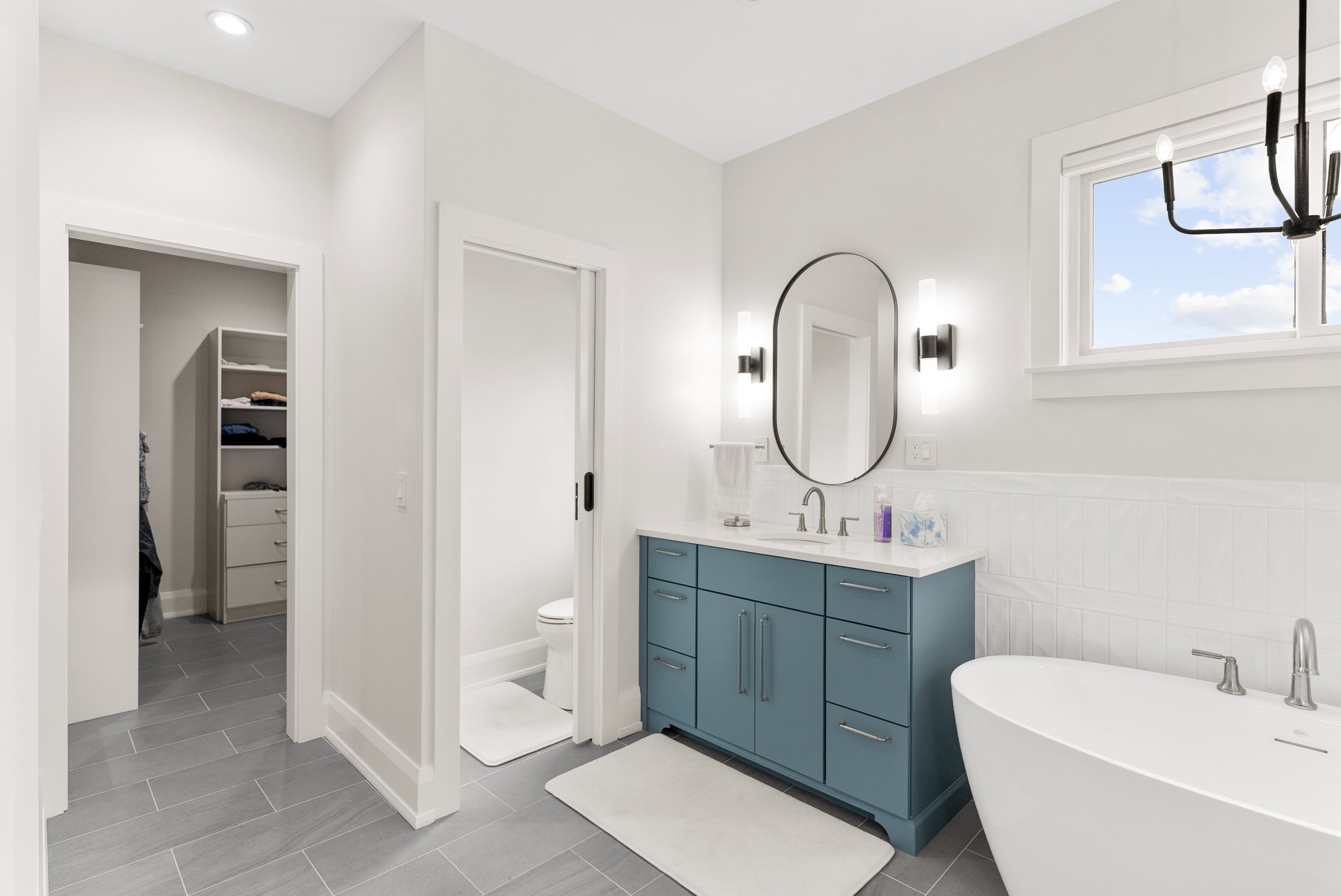 Modern bathroom with white walls, gray tile floor, blue vanity with white countertop, oval mirror, black light fixtures, window with blue sky view, bathtub, and a small laundry nook with shelves.